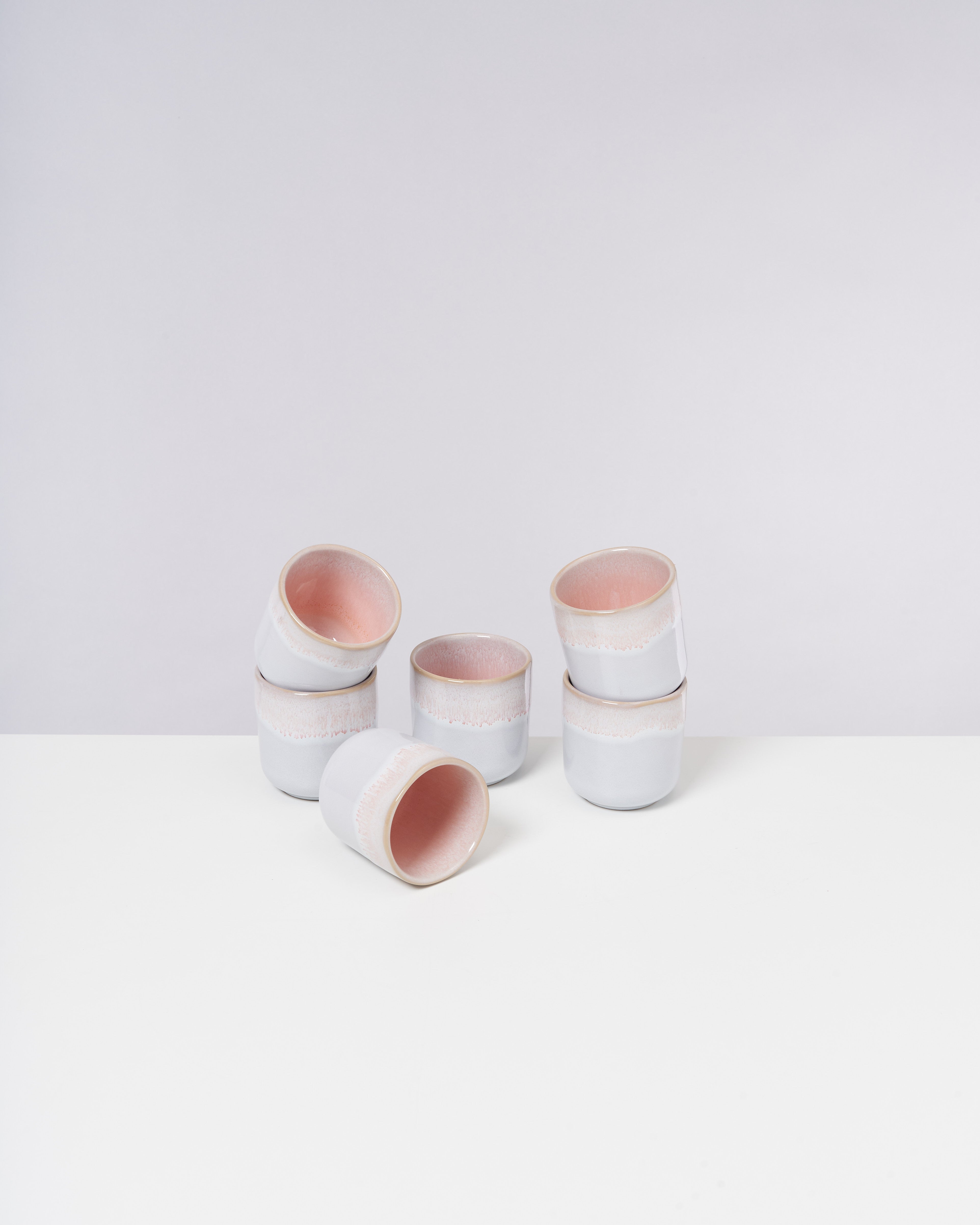 Sieben weiß-rosa Keramikbecher auf einem weißen Tisch vor einem hellgrauen Hintergrund.