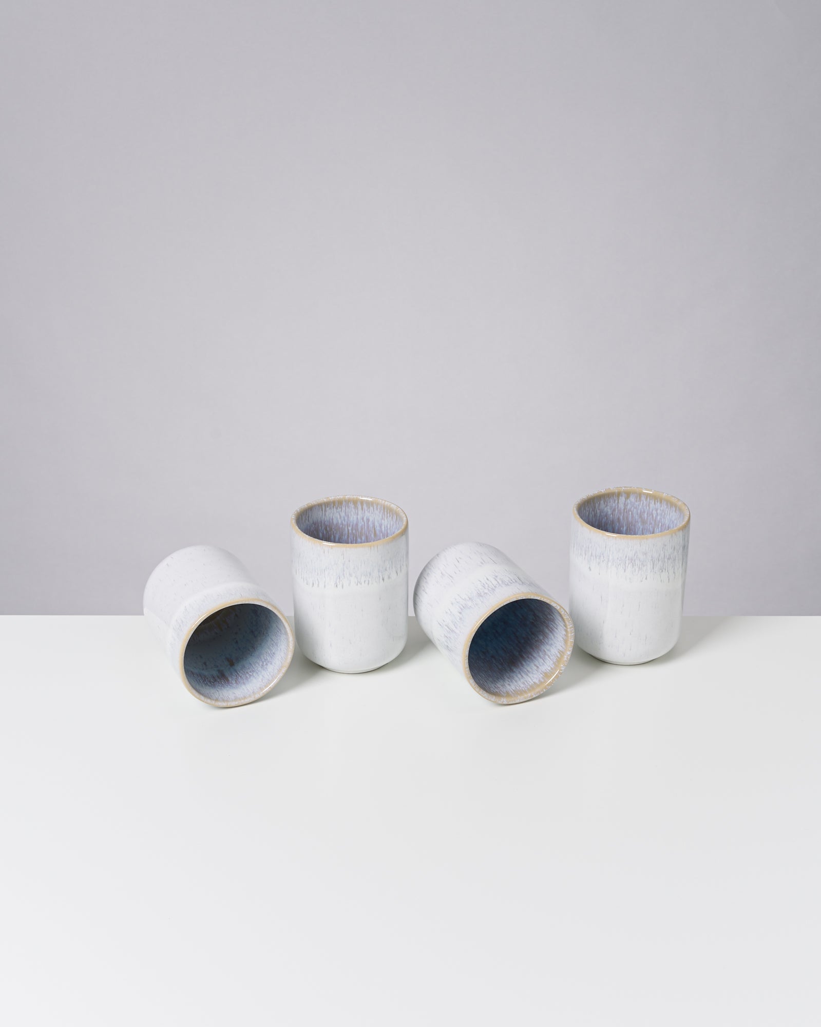 Vier weiße, strukturierte Becher stehen und liegen auf einem weißen Tisch vor einem grauen Hintergrund.