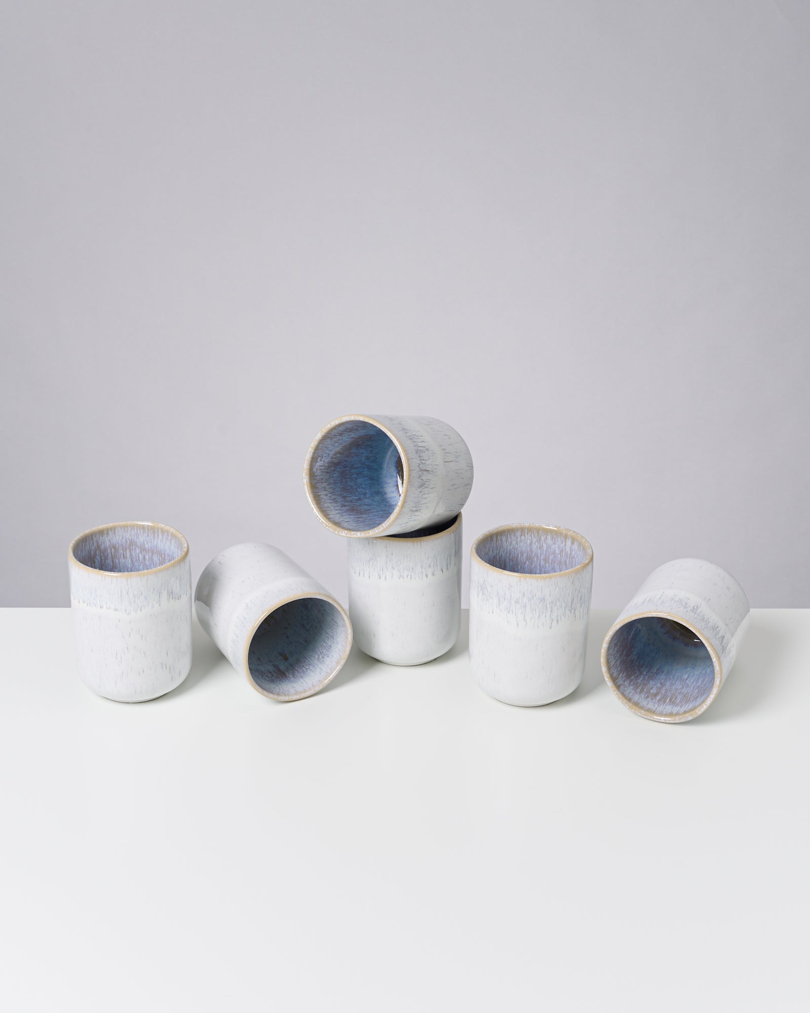 Fünf weiße Keramikbecher mit blauer Innenseite stehen auf einem weißen Tisch vor einem grauen Hintergrund.