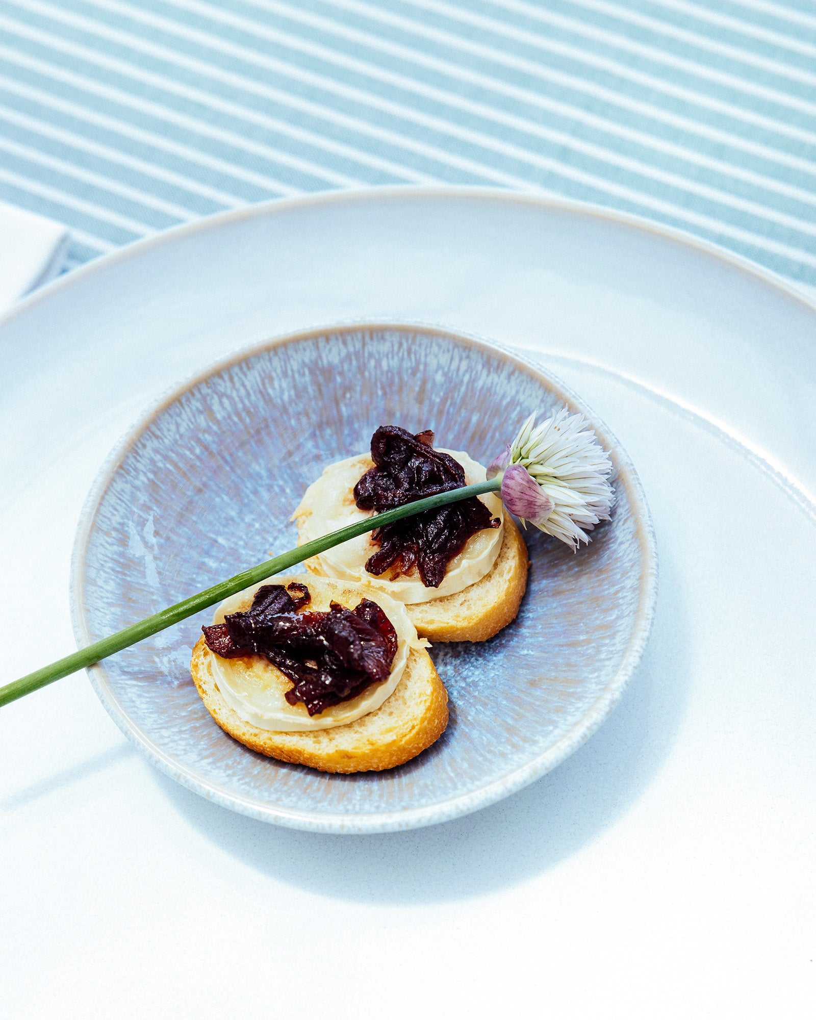 Kleine Brotscheiben mit Ziegenkäse und Zwiebelmarmelade auf einem blauen Teller, dekoriert mit einer Blume.