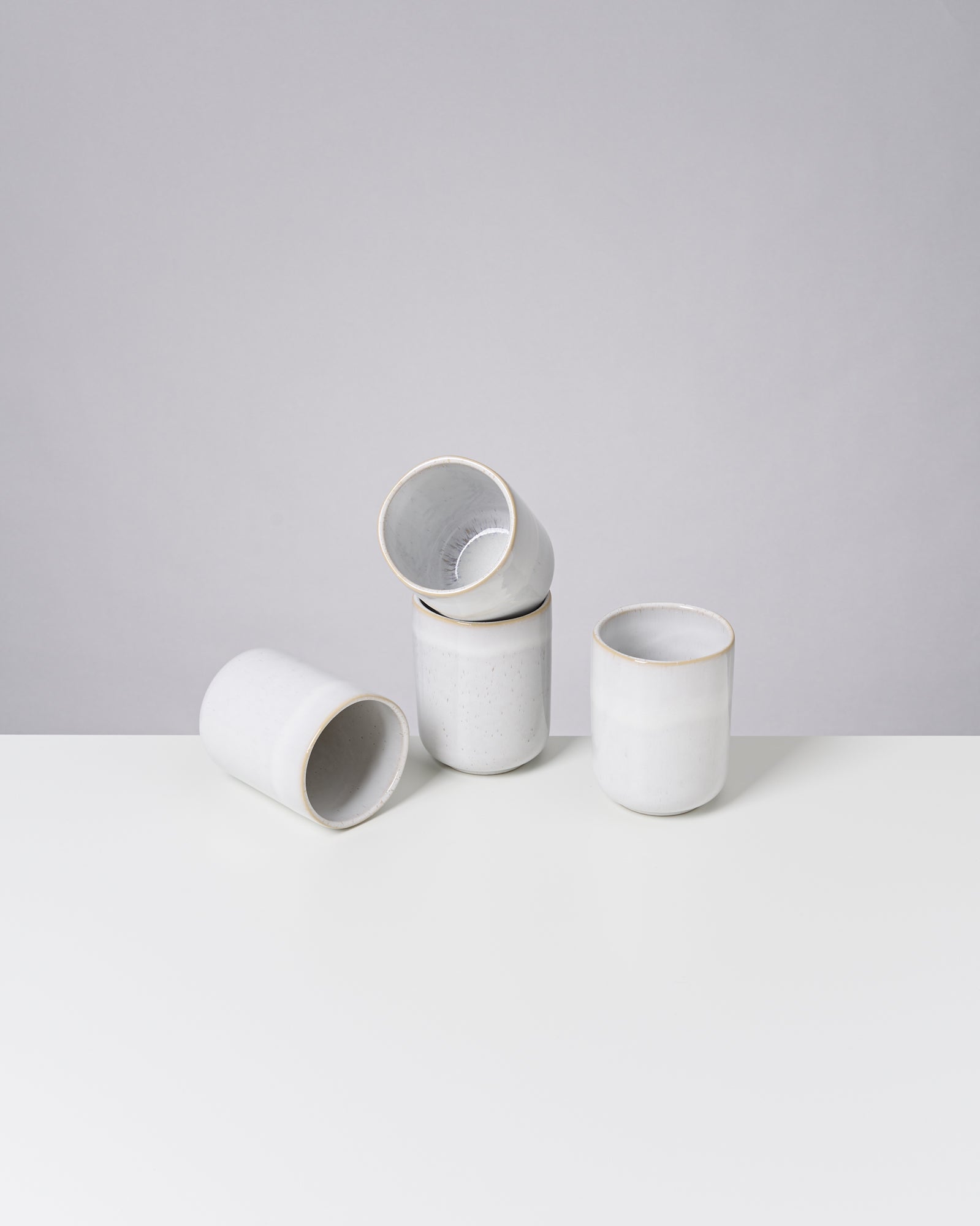 Vier weiße, minimalistische Keramiktassen auf einem weißen Tisch vor einem grauen Hintergrund.