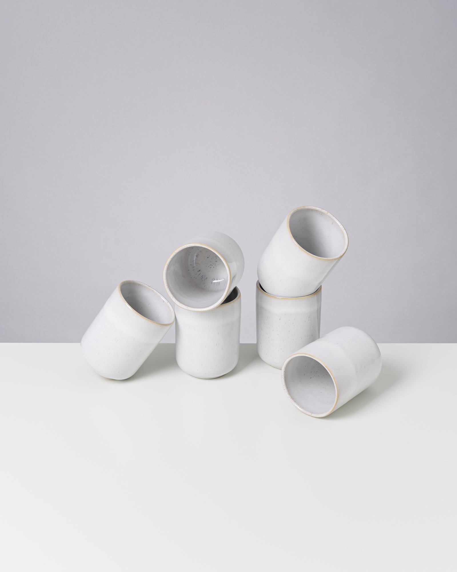Sechs weiße, zylindrische Keramiktassen auf einem weißen Tisch vor einer grauen Wand, in einem unregelmäßigen Arrangement.