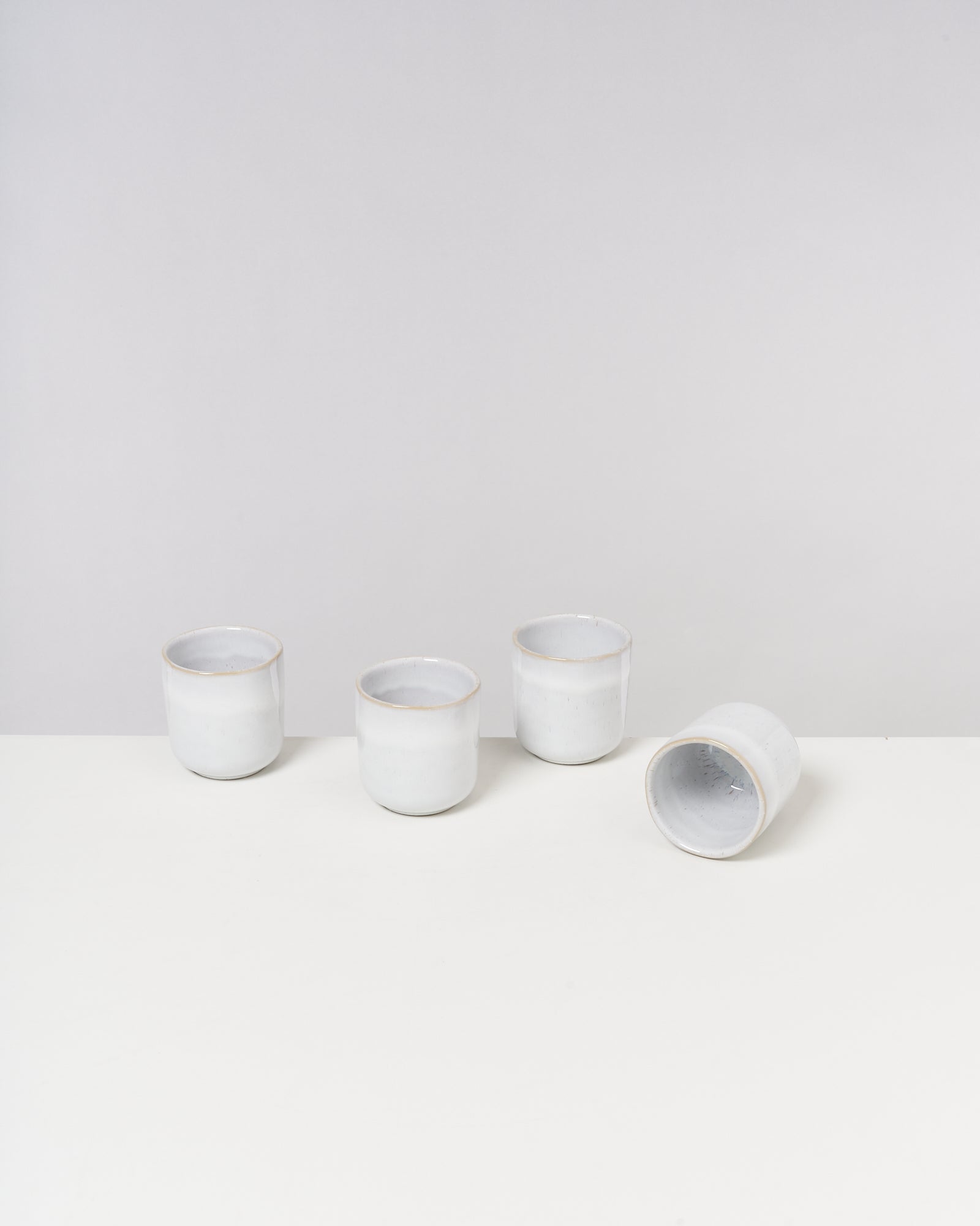 Vier weiße, keramikähnliche Becher auf einem weißen Tisch, einer liegt seitlich.