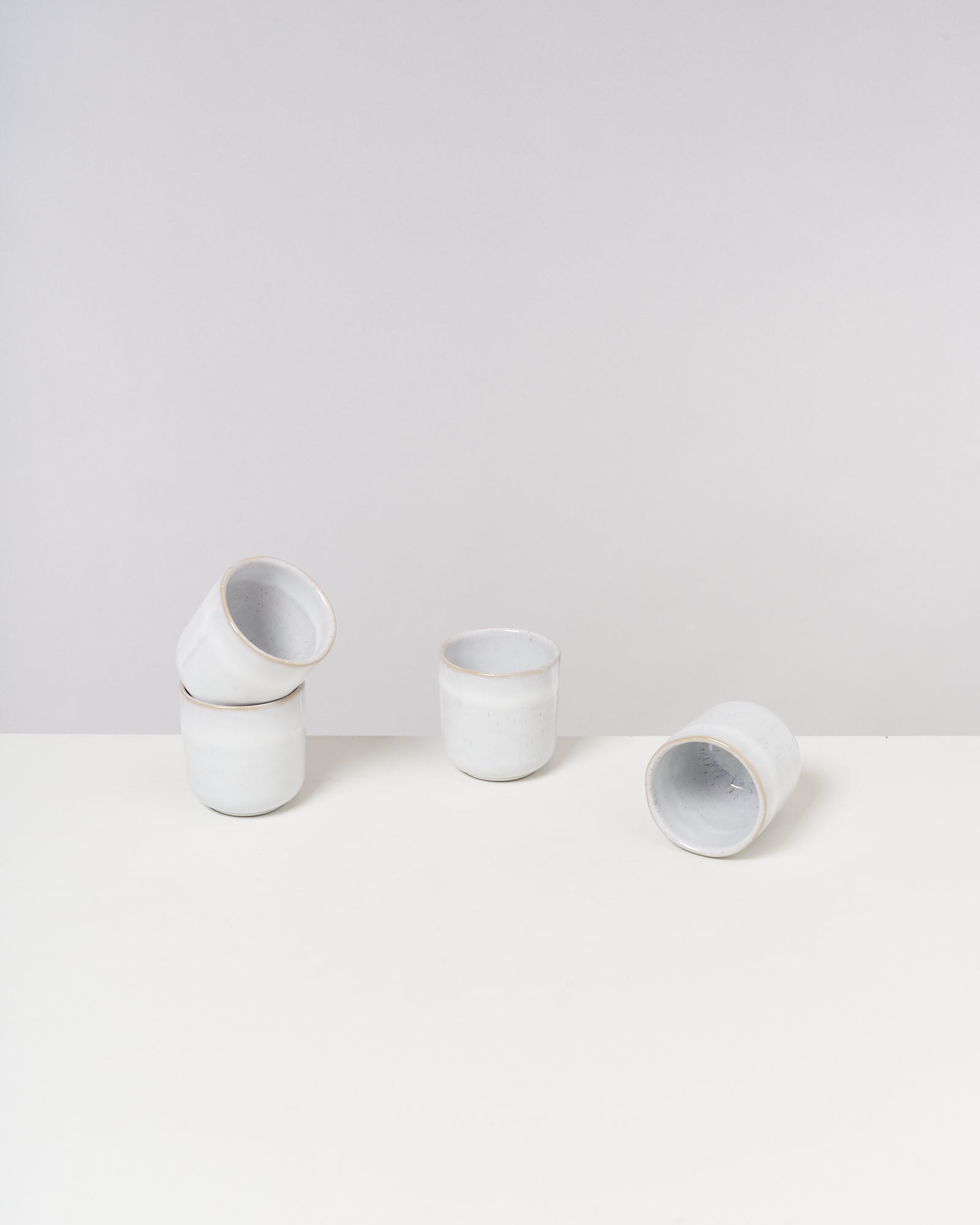 Vier weiße Keramiktassen, zwei gestapelt, eine stehend, eine liegend, vor grauem Hintergrund.