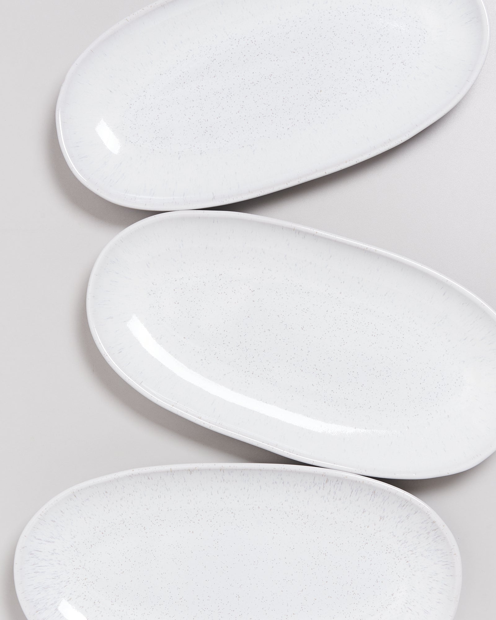 Drei weiße, ovale Teller mit dezentem Sprenkelmuster liegen auf einer grauen Fläche.