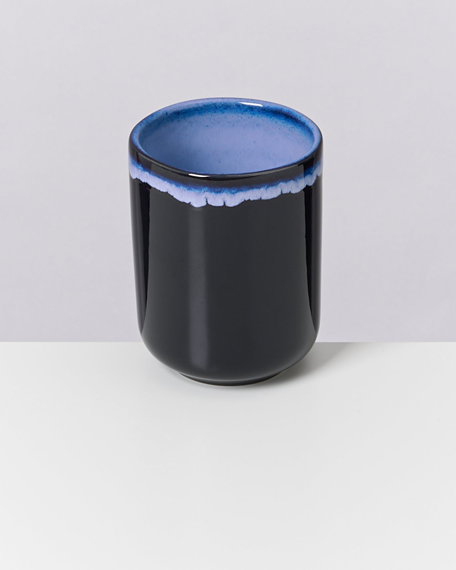 Schwarzer Keramikbecher mit blauem Rand auf einem weißen Tisch vor grauem Hintergrund.