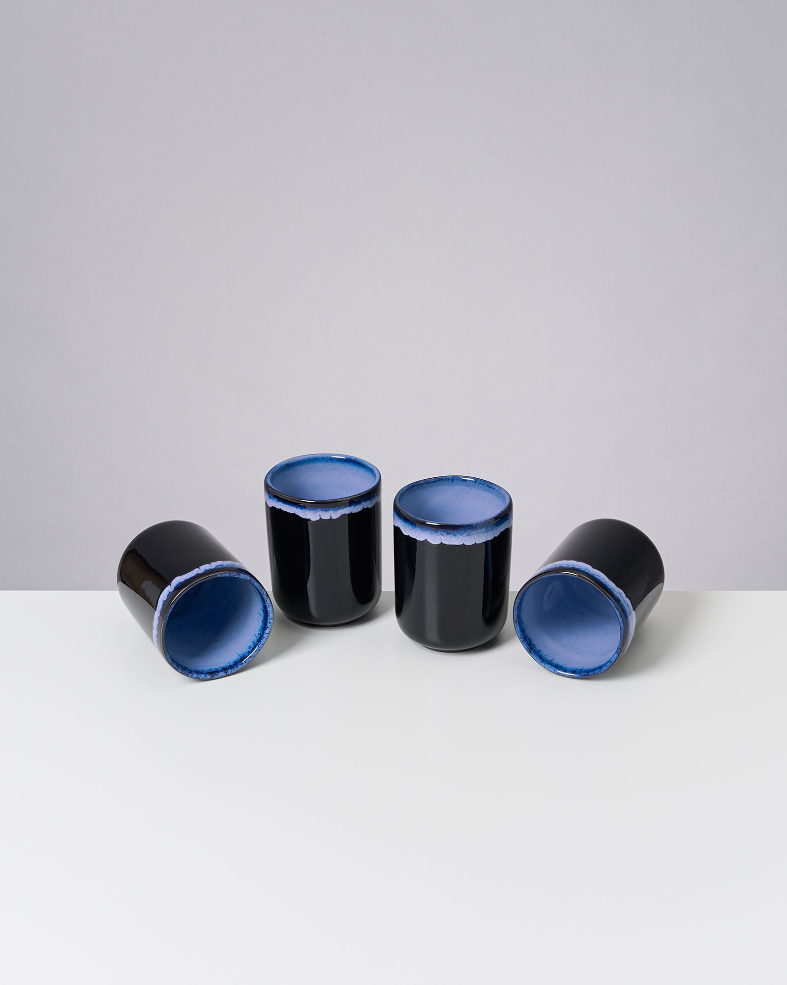Vier schwarze Becher mit blauen Innenwänden auf einem weißen Tisch vor grauem Hintergrund.