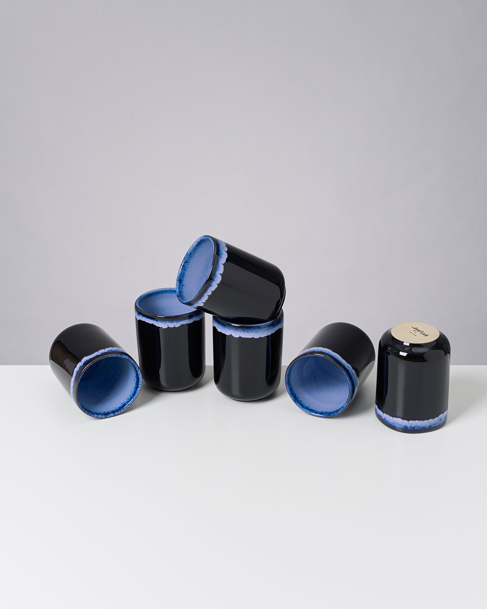 Sechs schwarze Tassen mit blauer Innenseite, teils gestapelt, auf einem weißen Tisch vor grauem Hintergrund.