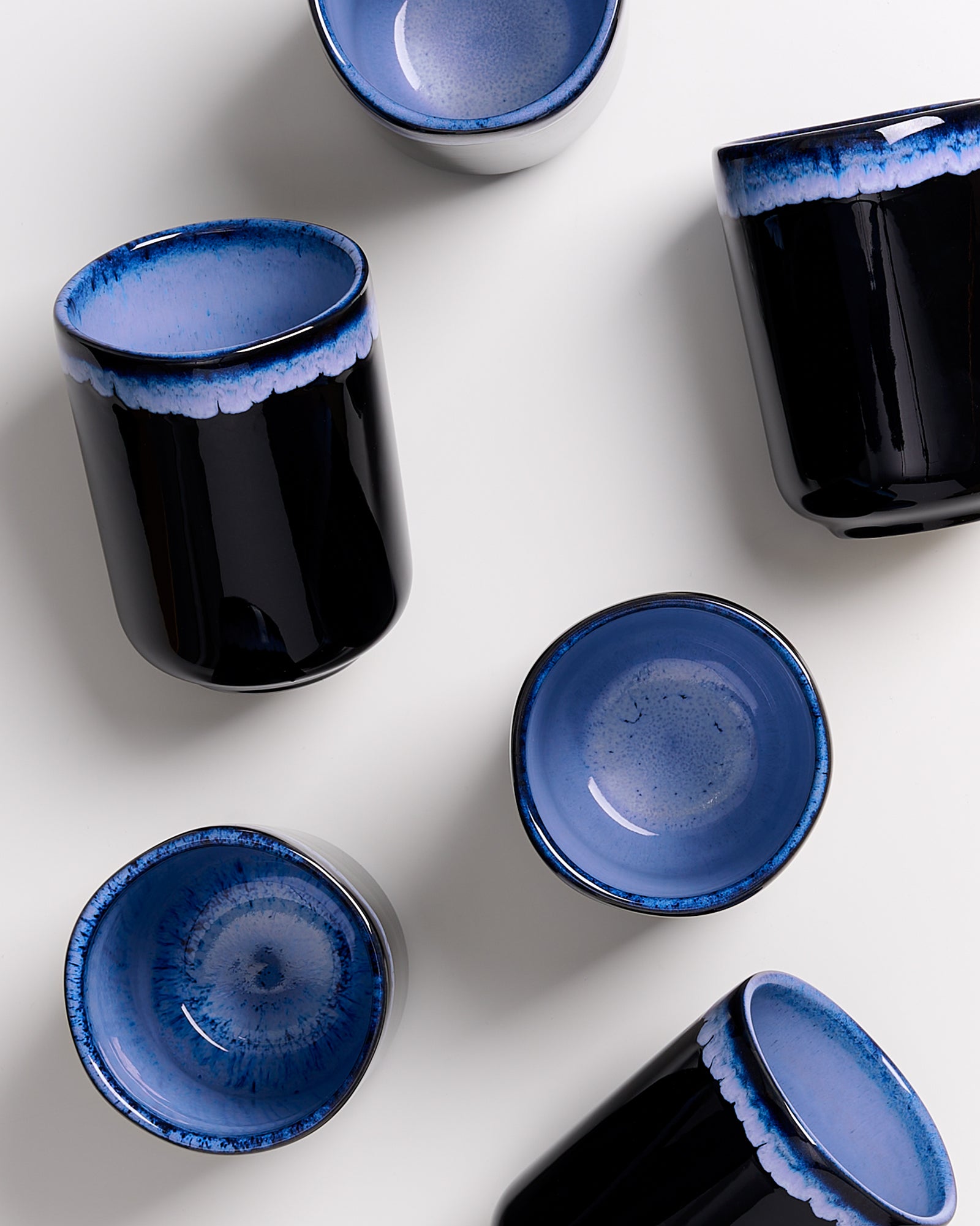 Sechs blau-schwarze, glasierte Keramiktassen liegen auf einem weißen Hintergrund verstreut.