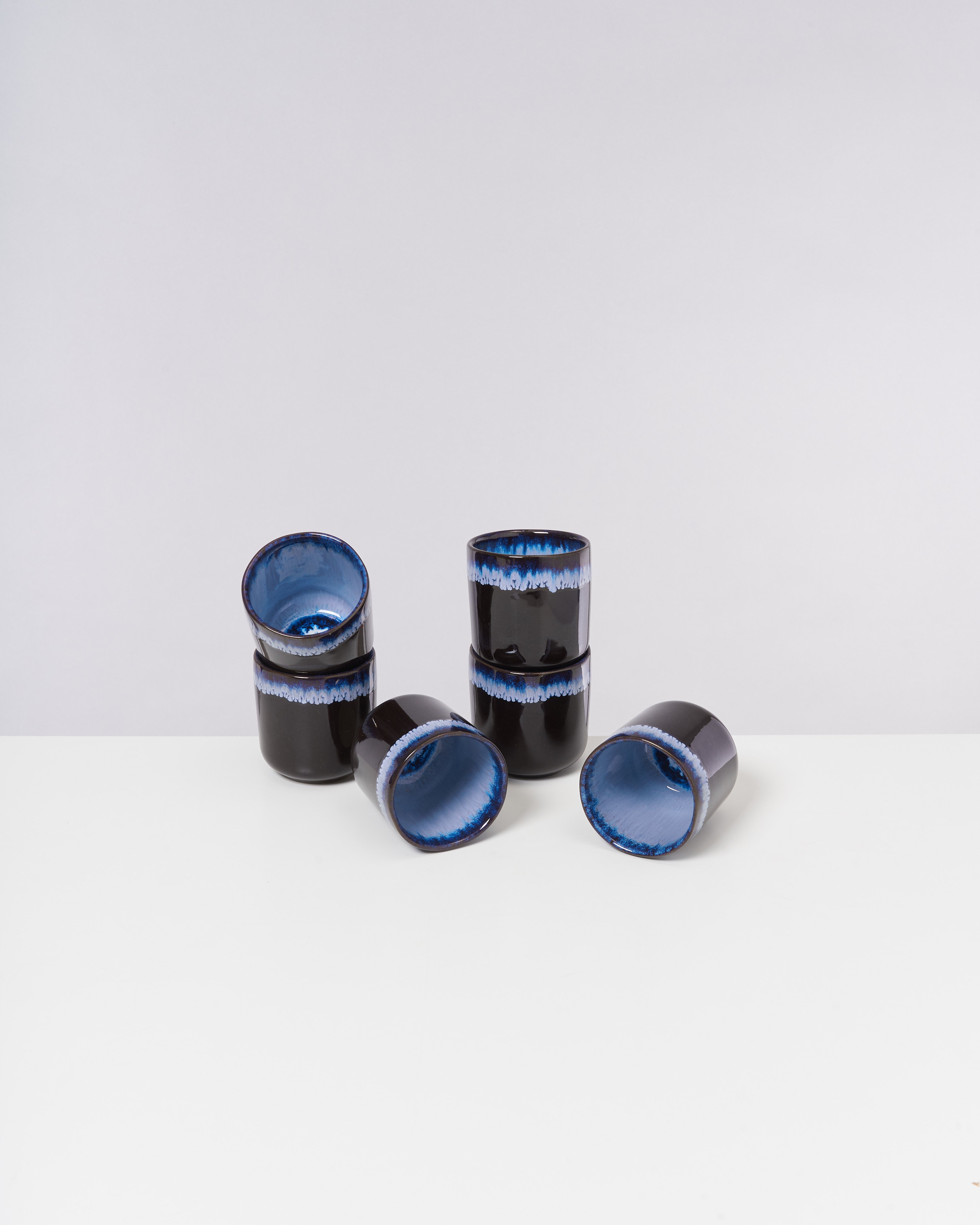 Sechs schwarze Tassen mit blauer Innenverzierung stehen und liegen auf einem weißen Tisch.