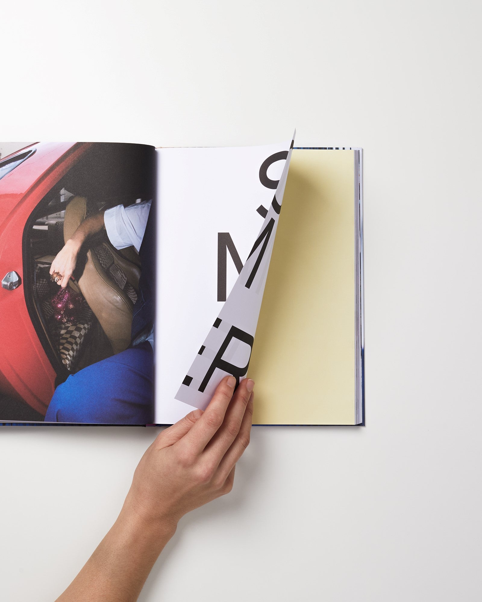Eine Hand blättert eine Buchseite um; links ist ein Autoausschnitt zu sehen.