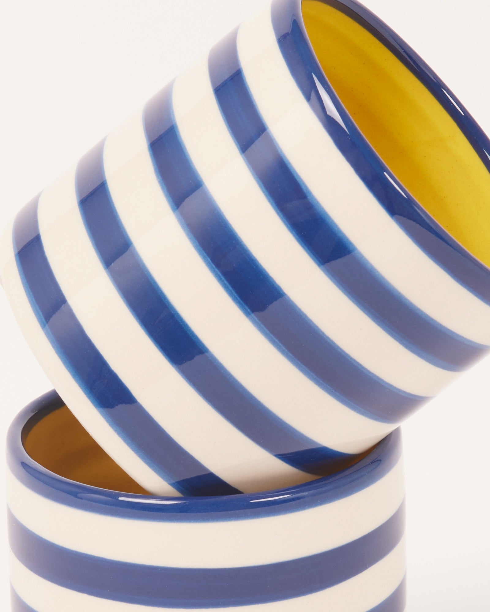 Gestapelte, blau-weiß gestreifte Keramikbecher mit gelber Innenseite vor weißem Hintergrund.