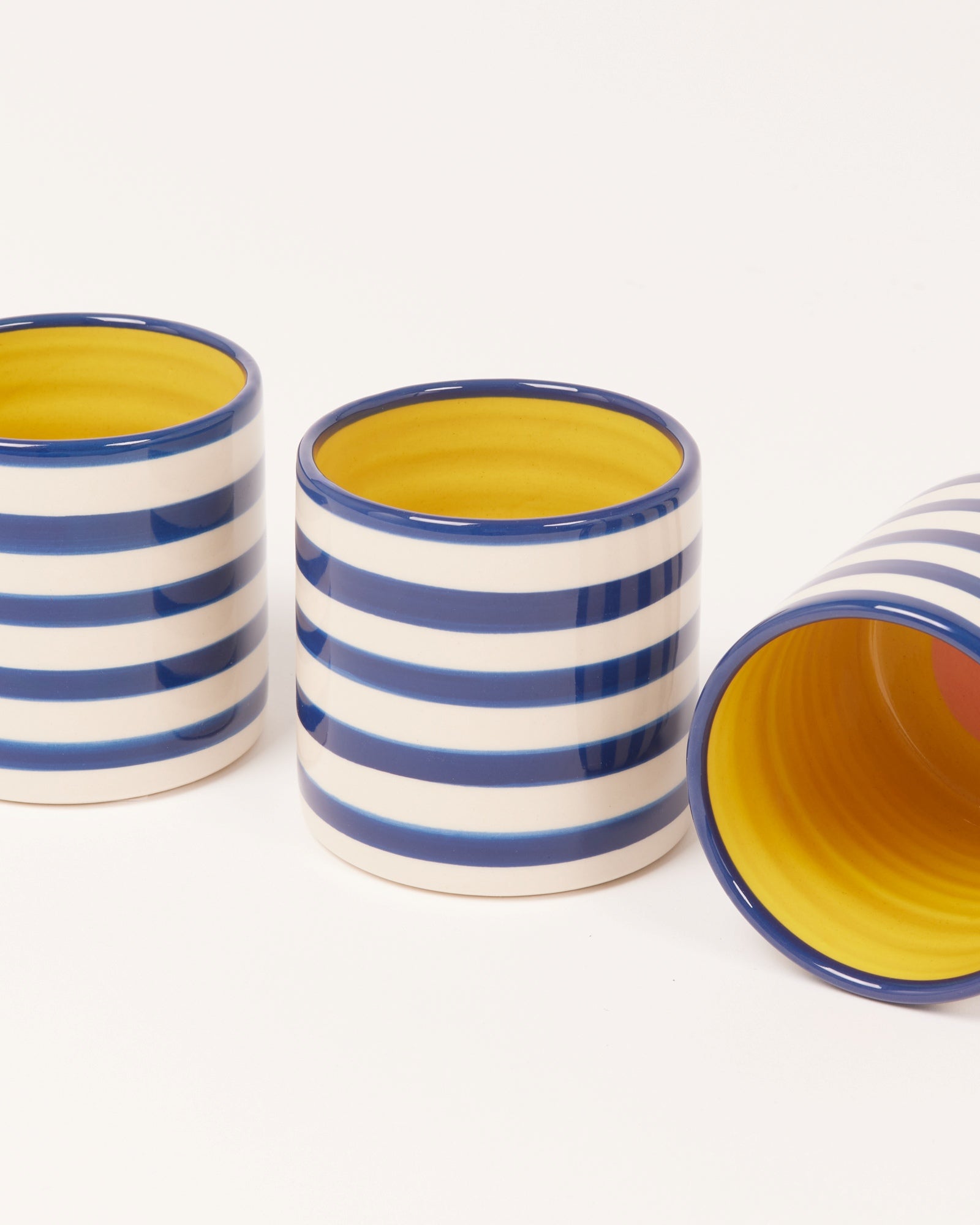 Drei Keramiktassen mit blauen und weißen Streifen, gelb im Inneren. Eine liegt seitlich auf dem weißen Hintergrund.
