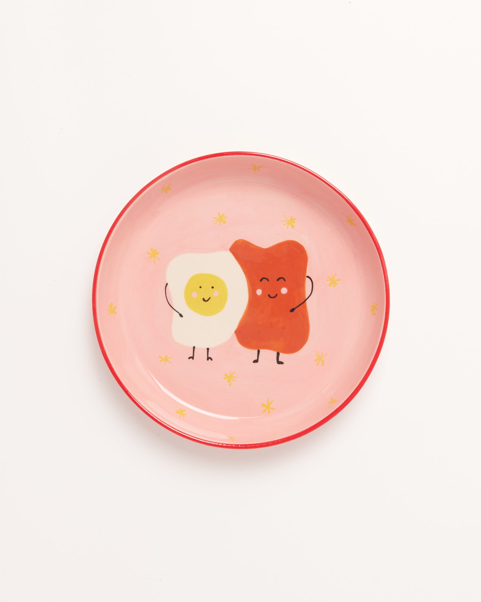 Kreisförmiger Teller mit einem gemalten, lächelnden Spiegelei und Toast auf rosa Hintergrund mit gelben Sternen.