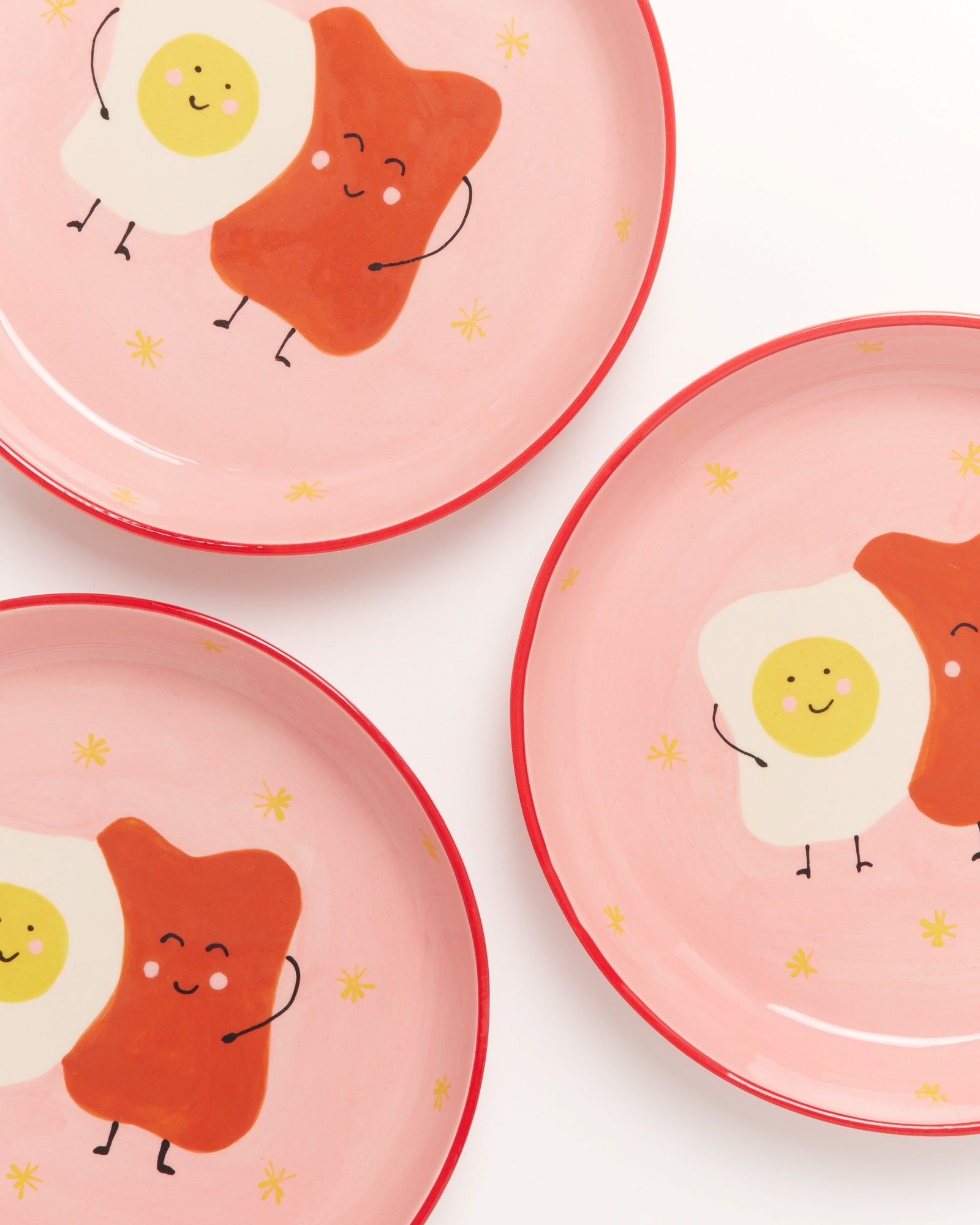 Drei rosa Teller mit niedlichen Illustrationen von Spiegelei und Toast, die sich umarmen, auf weißem Hintergrund.