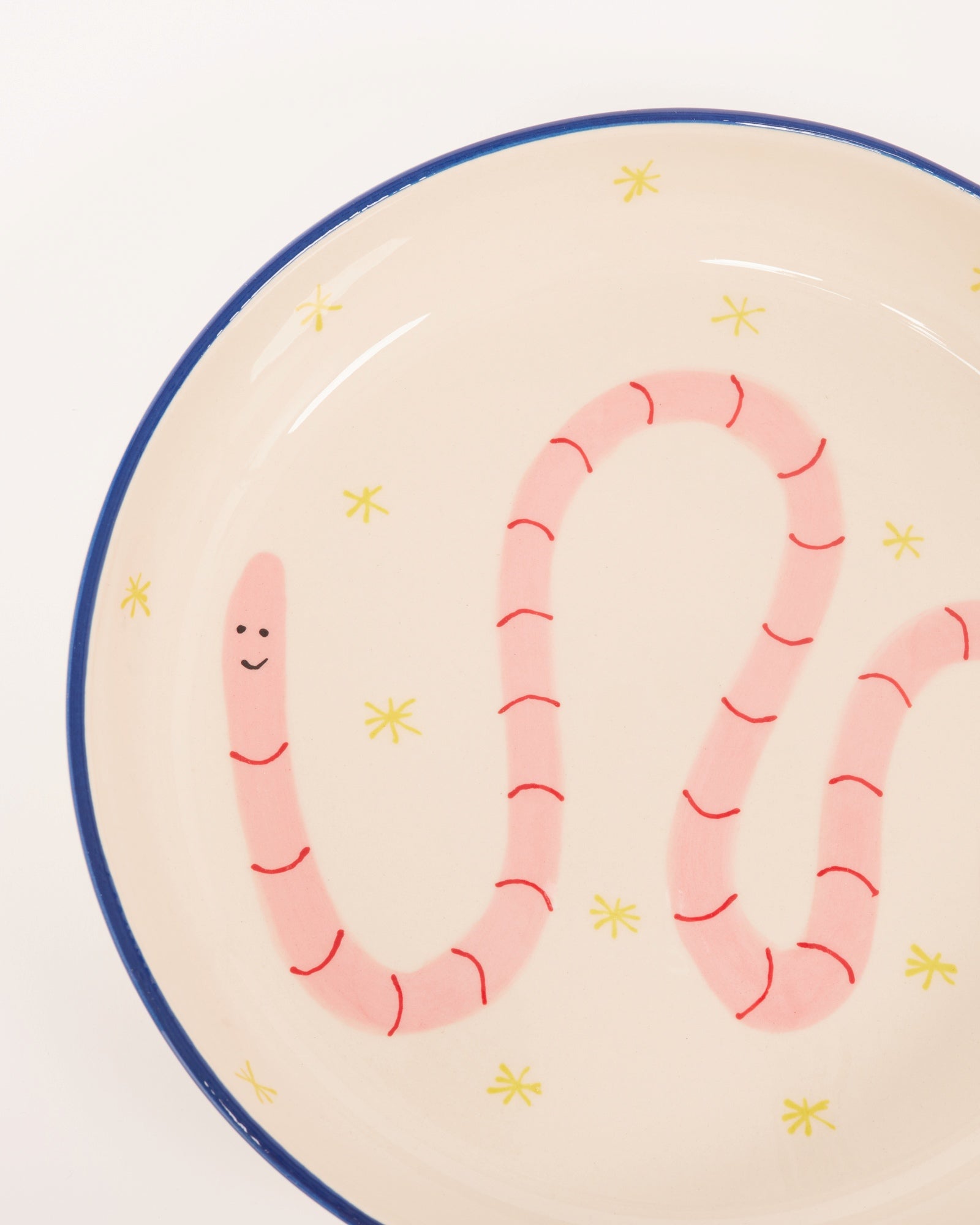 Keramikschüssel mit blauem Rand, bemalt mit rosa Wurm und gelben Sternen auf cremefarbenem Hintergrund.