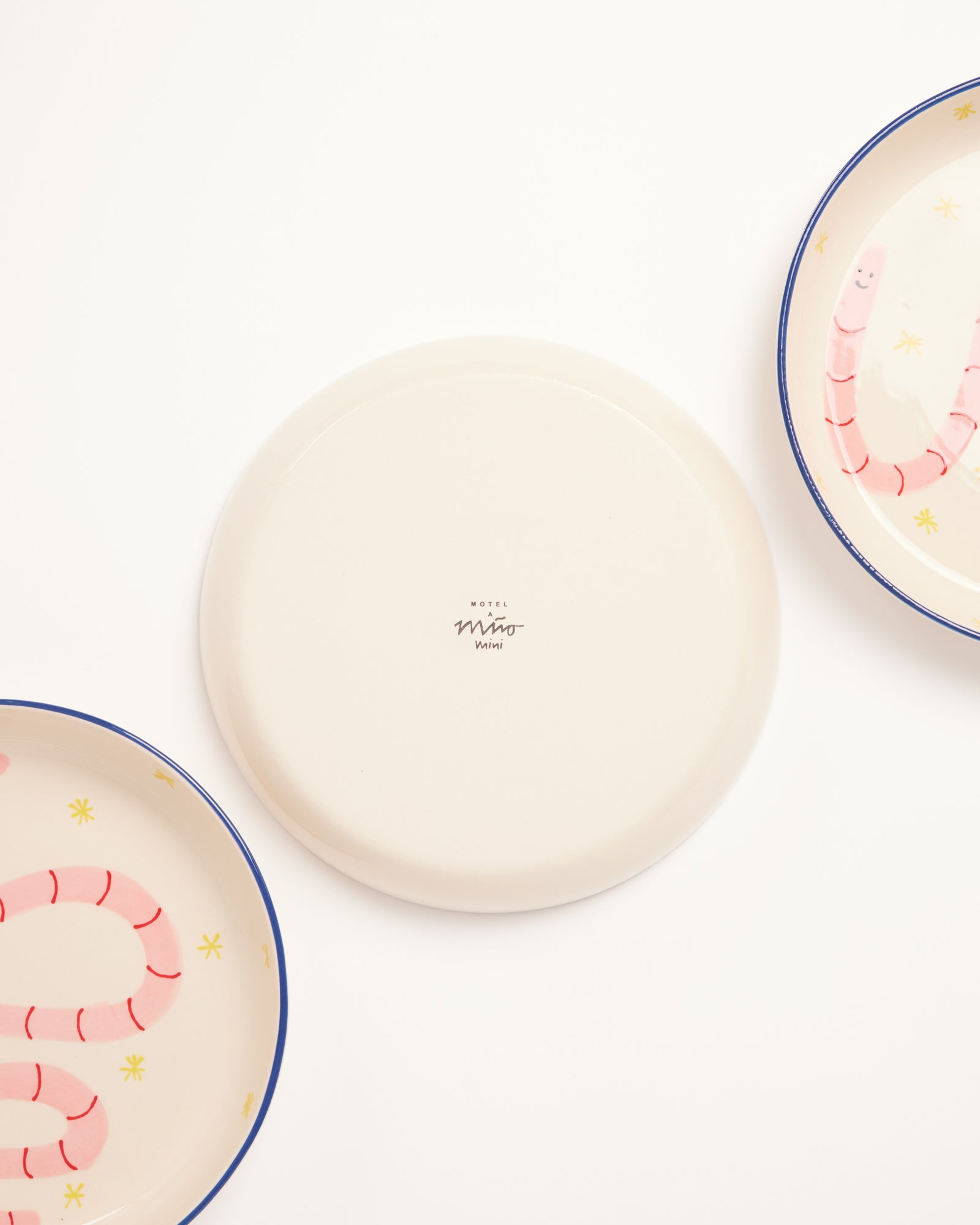Drei Teller, zwei davon mit rosa Würmchenmuster, der mittlere zeigt die Unterseite mit der Aufschrift "Motel Miro Waik".