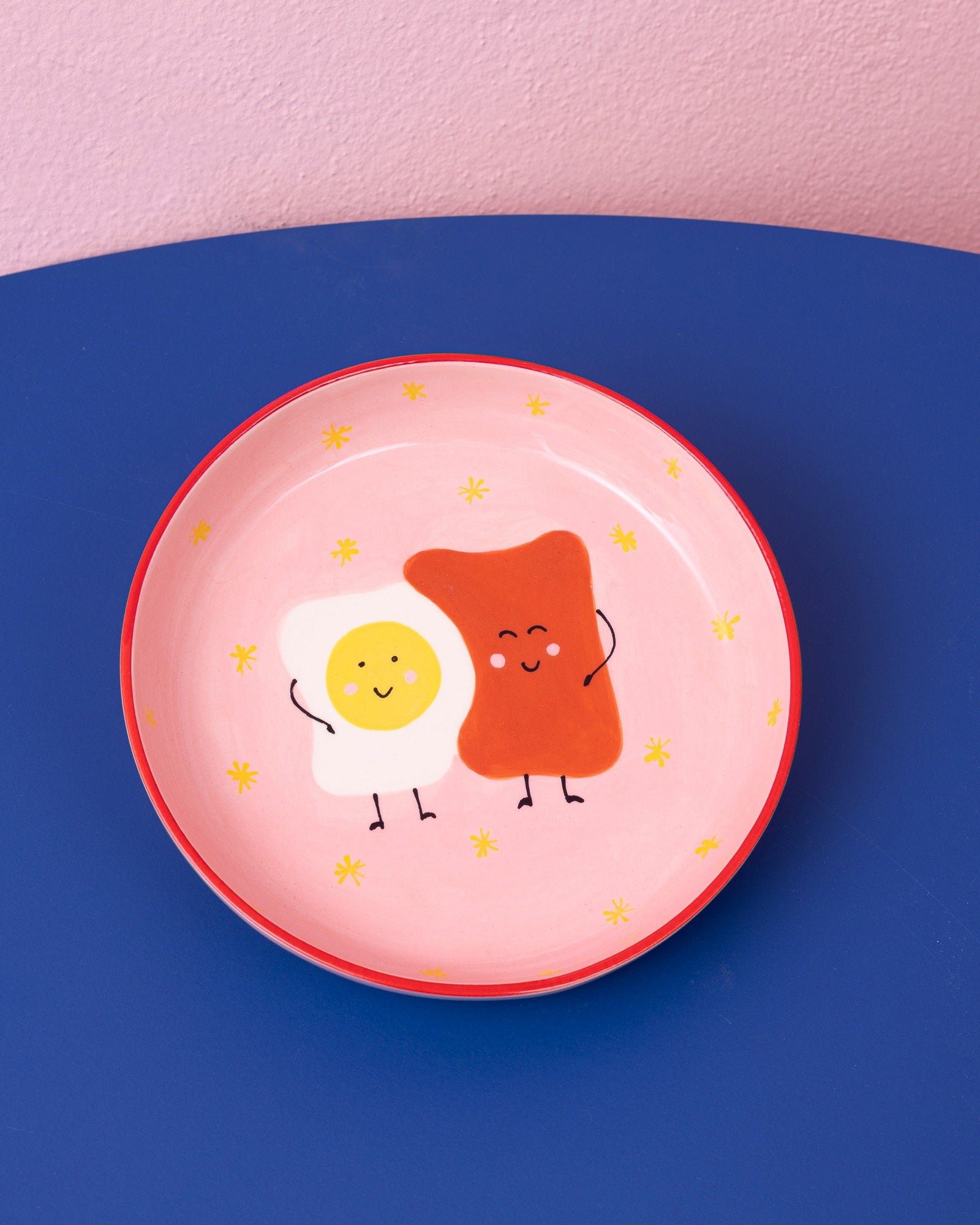 Teller mit einem lächelnden, gezeichneten Spiegelei und Toast auf rosa Hintergrund mit gelben Sternen.