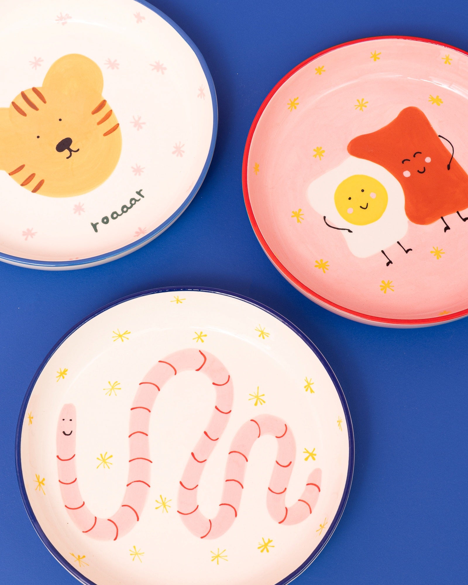 Drei bunte Teller mit niedlichen Illustrationen, darunter ein Tiger, ein Wurm und ein lächelndes Ei mit Toast.