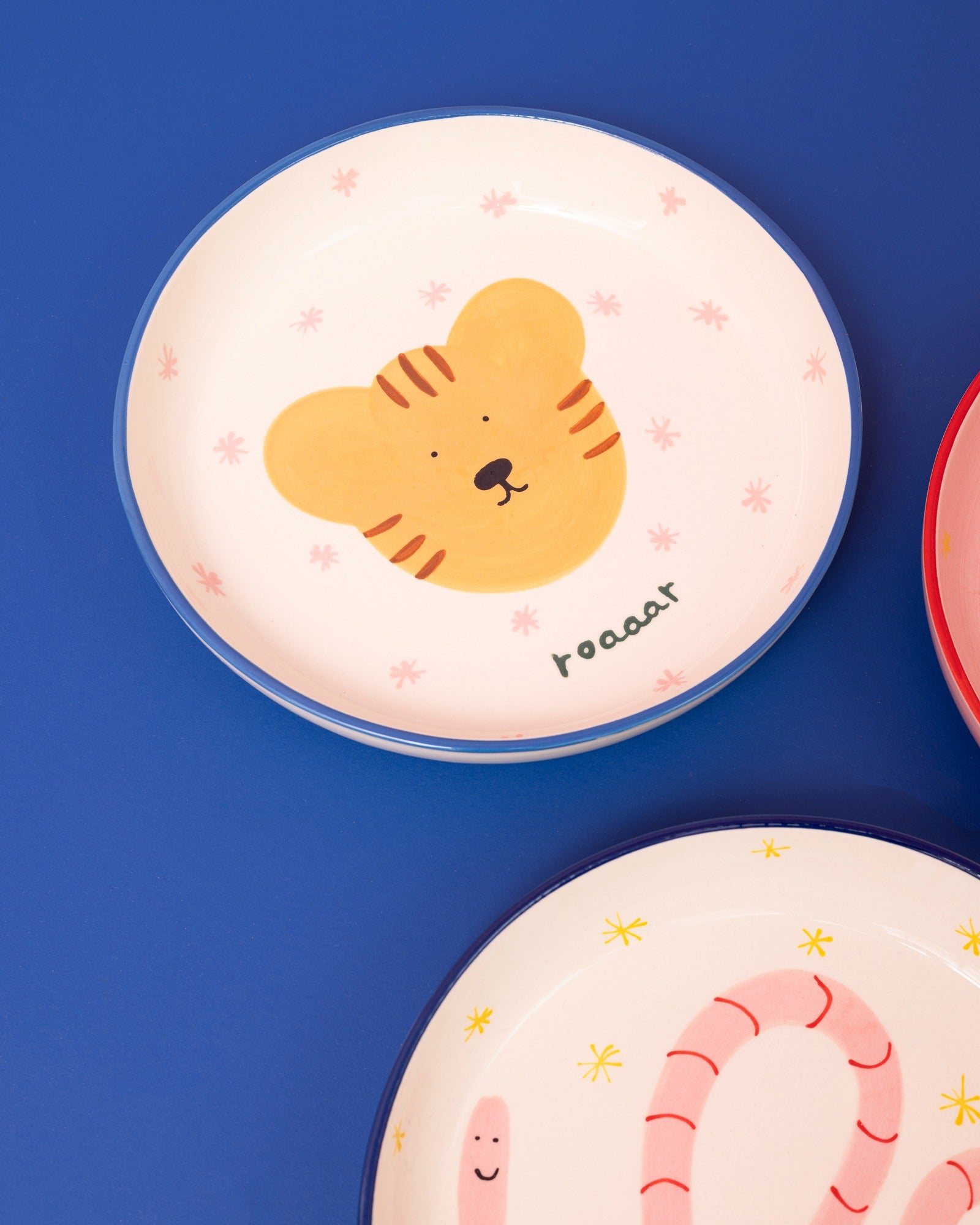 Zwei dekorative Teller auf blauem Hintergrund, einer mit einem Tigerkopf-Motiv und der andere mit bunten Mustern.