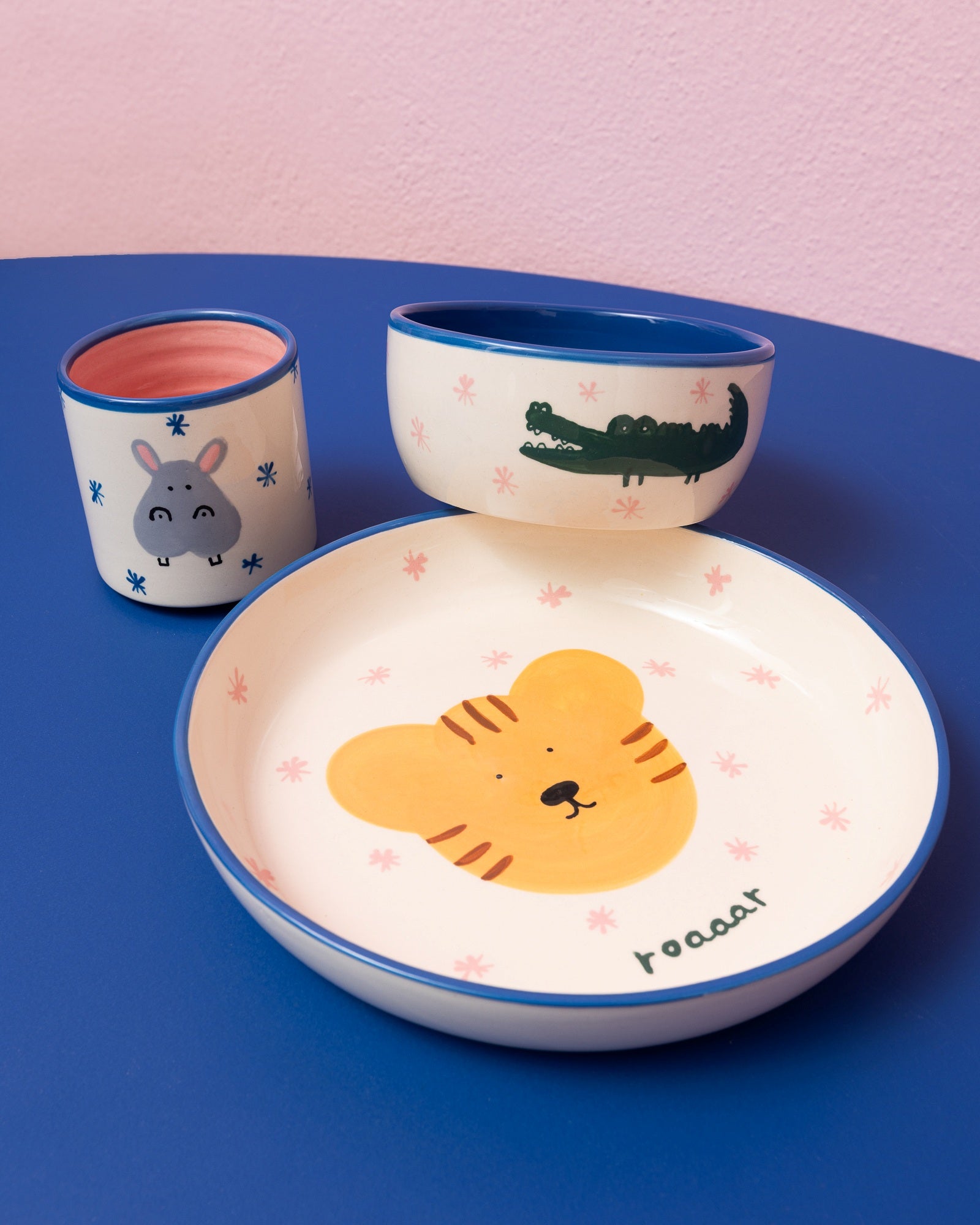 Kindergeschirr-Set mit Tiermotiven: Teller mit Tiger, Schale mit Krokodil und Becher mit Nilpferd auf blauem Tisch.