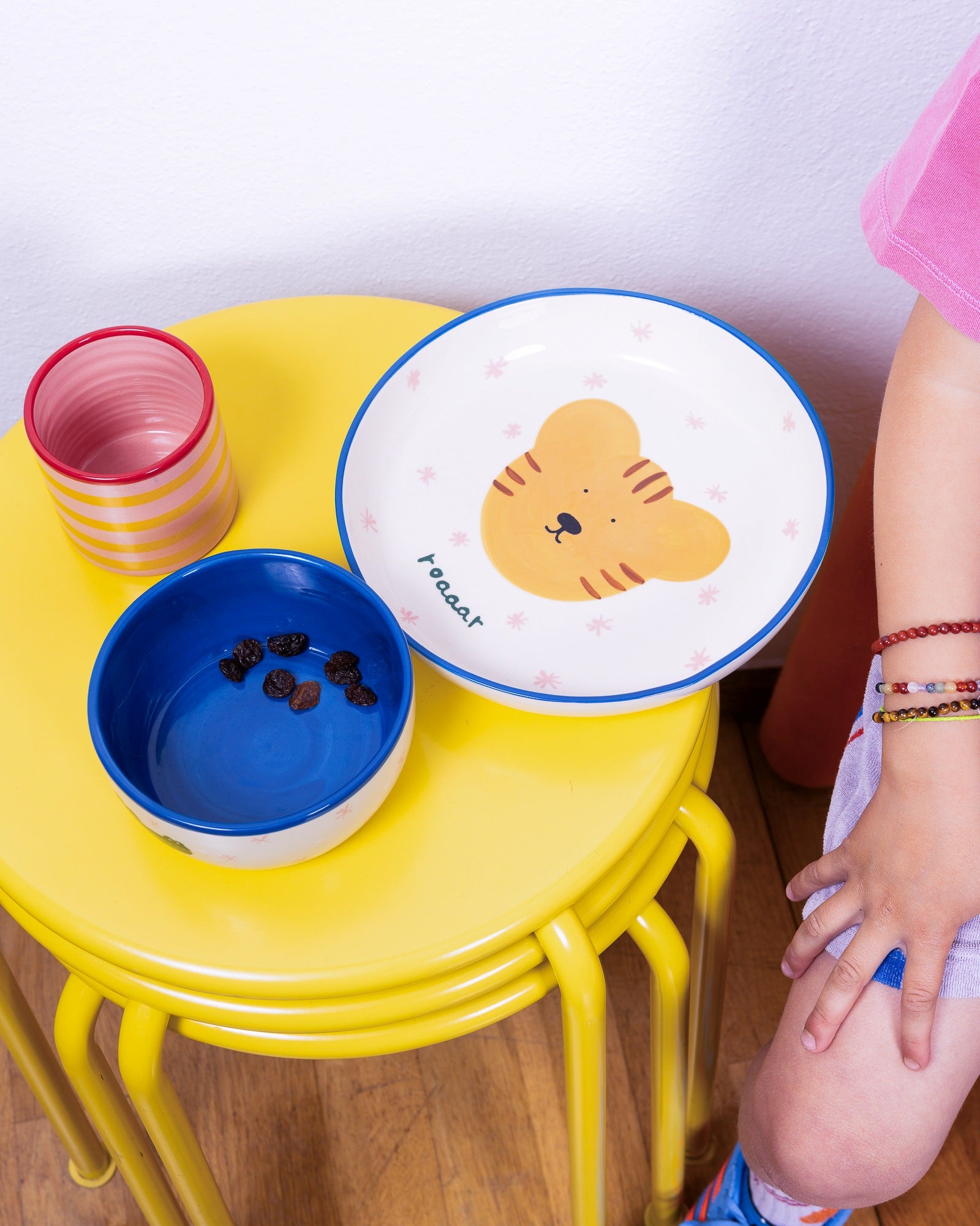 Ein gelber Tisch mit Schüsseln, eine mit Tigermotiv, die andere mit Rosinen. Ein Kind in rosa Kleidung daneben.