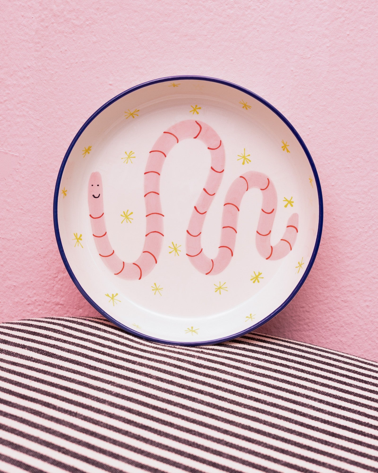 Teller mit rotem Wurm-Motiv und gelben Sternen darauf, vor einer rosa Wand auf gestreiftem Stoff platziert.