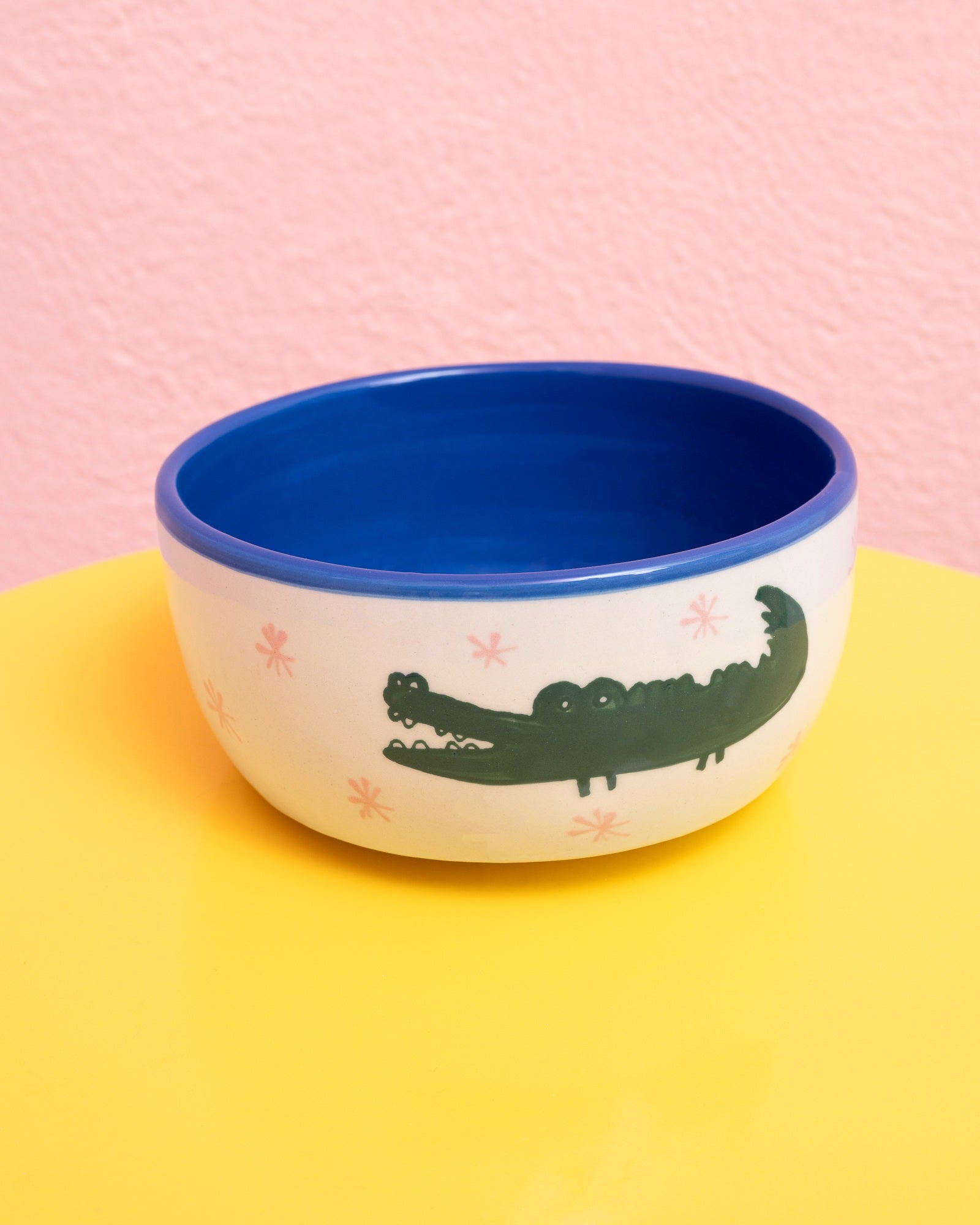 Weiße Schale mit Krokodilmuster und blauem Rand auf gelbem Tisch vor rosa Wand.