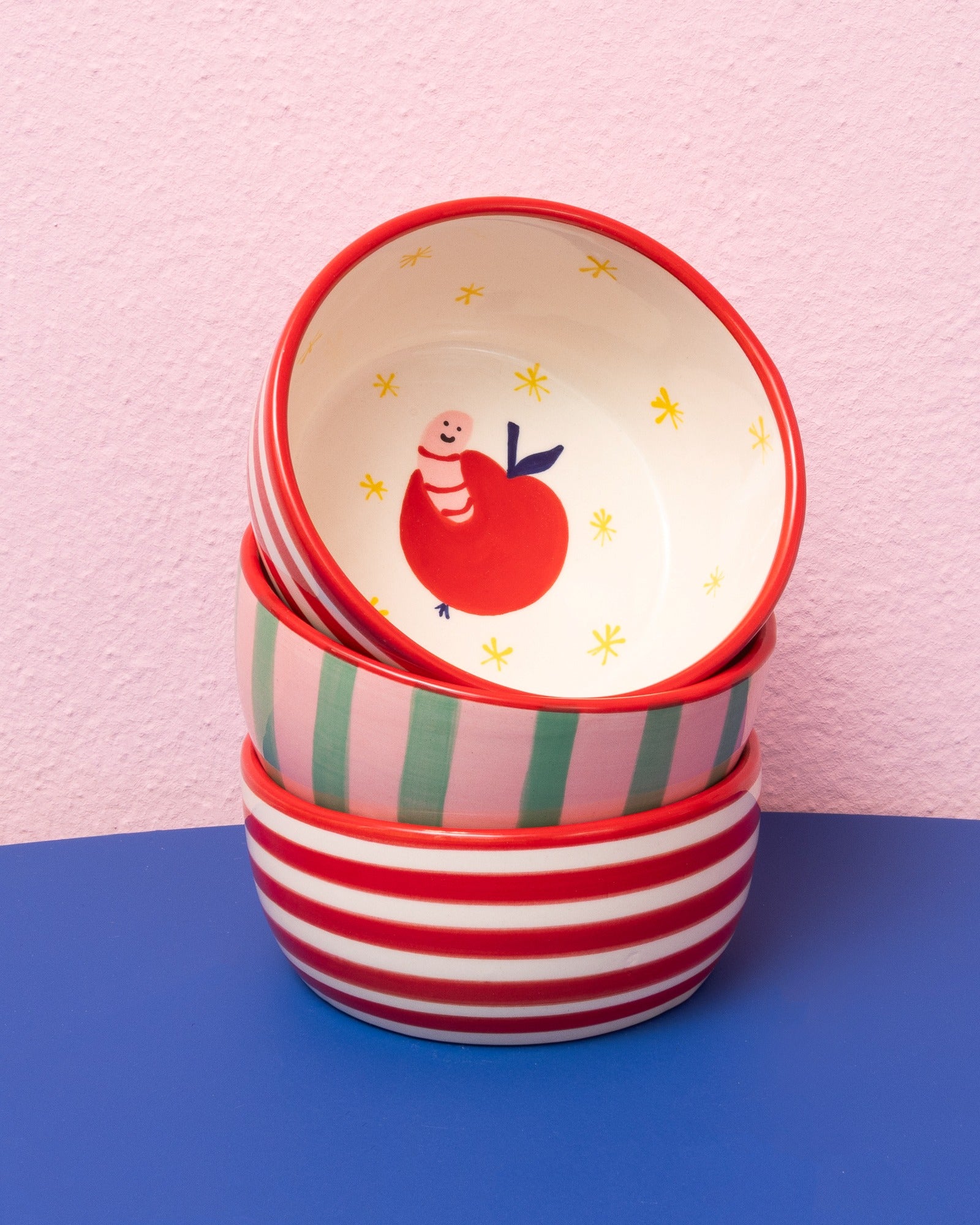 Vier bunte Schalen mit unterschiedlichen Designs stapeln sich vor einem rosa Hintergrund auf einer blauen Fläche.