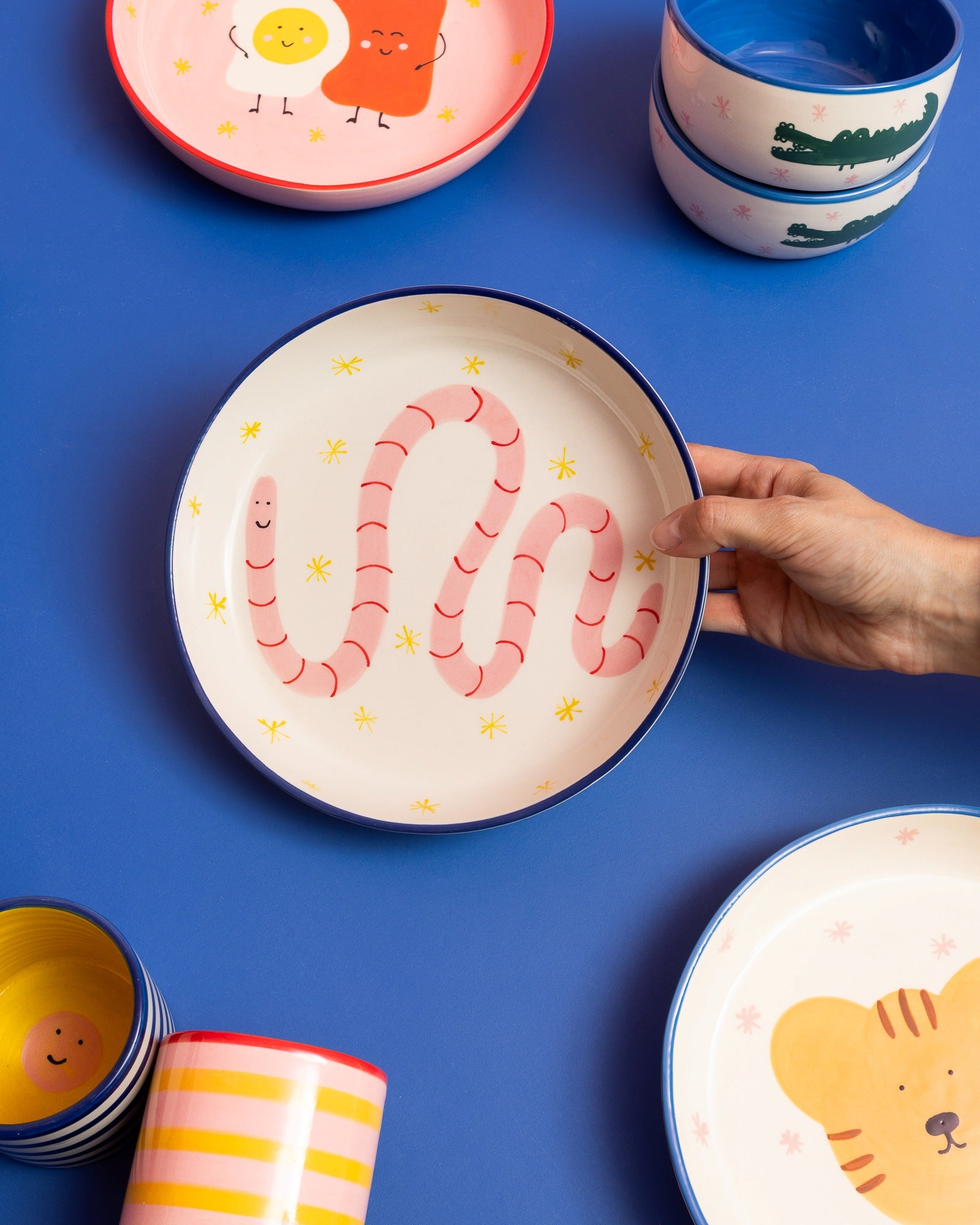 Eine Hand hält einen Teller mit einem rosa Wurm-Design, umgeben von weiteren Tellern und Schalen auf blauem Hintergrund.