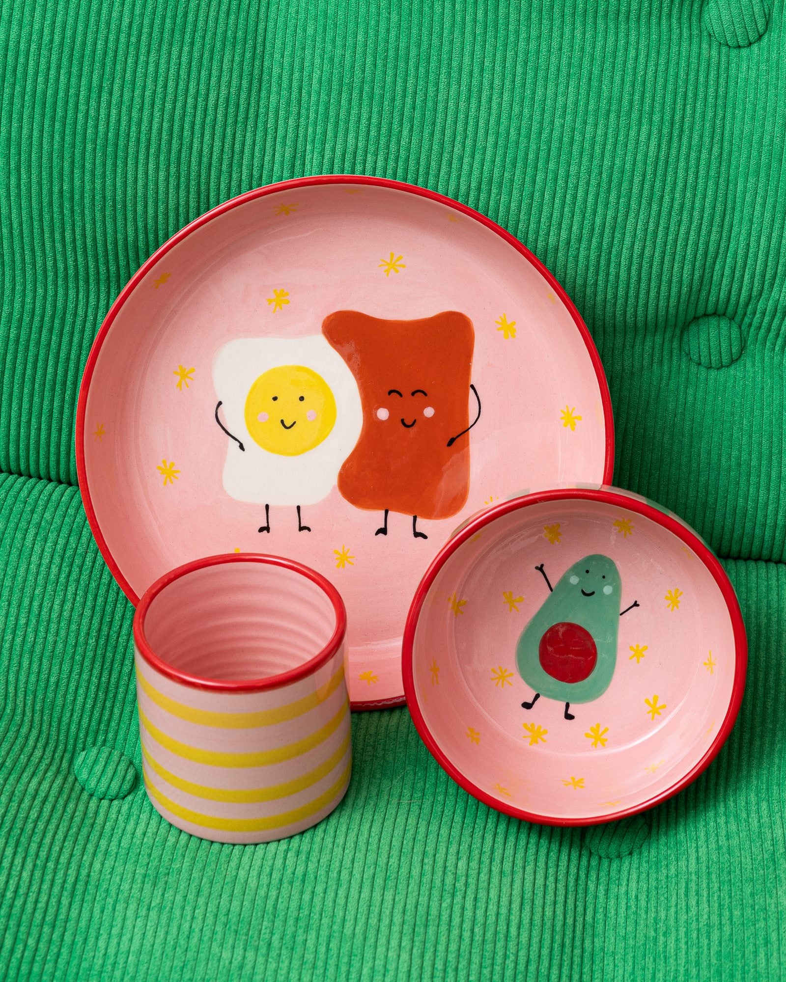 Drei rosa Geschirrteile mit fröhlichen Zeichnungen von Essen vor einem grünen, gestreiften Hintergrund.