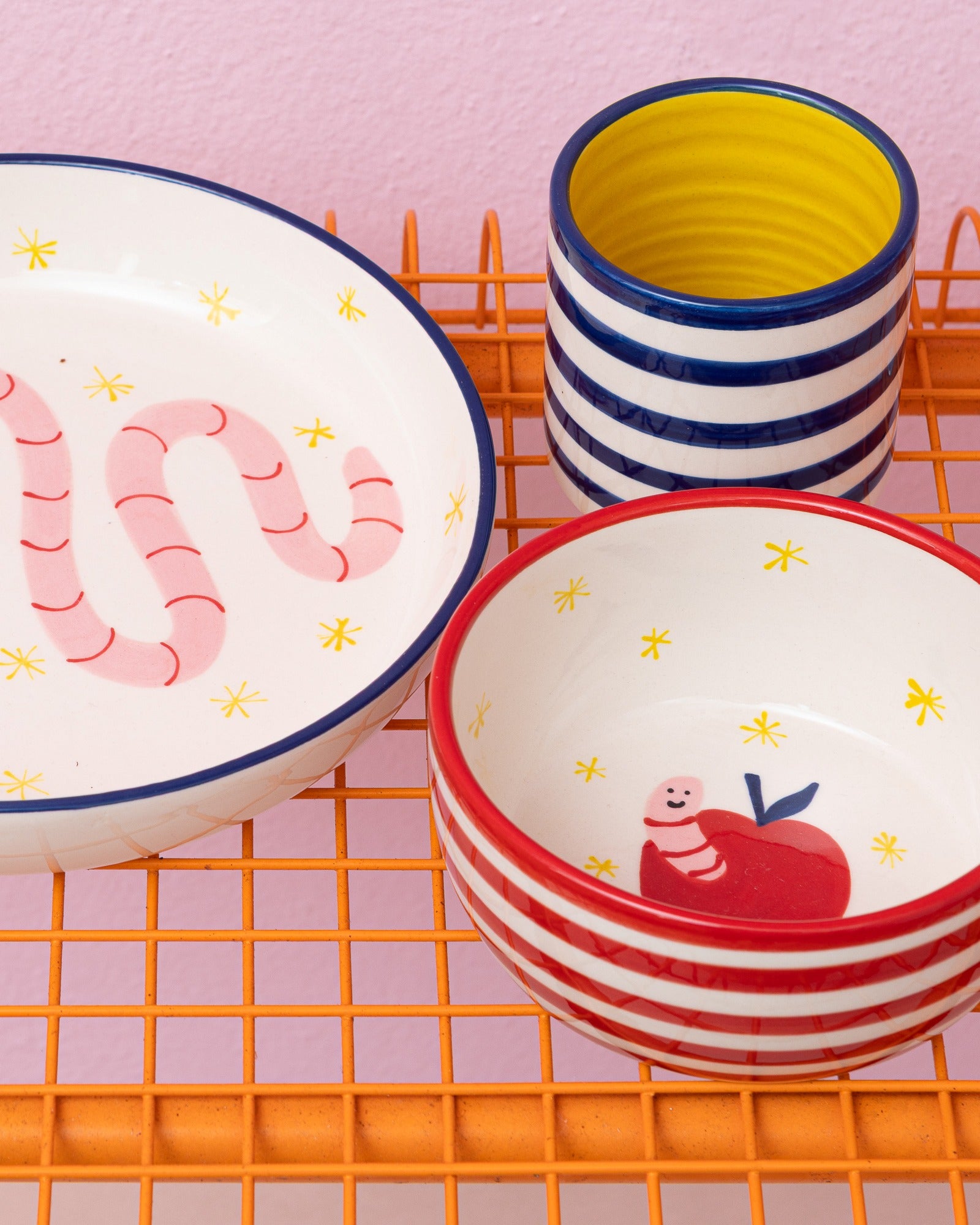 Bunte Schalen und Becher mit fröhlichen Designs auf einem orangefarbenen Drahtgestell vor rosa Hintergrund.
