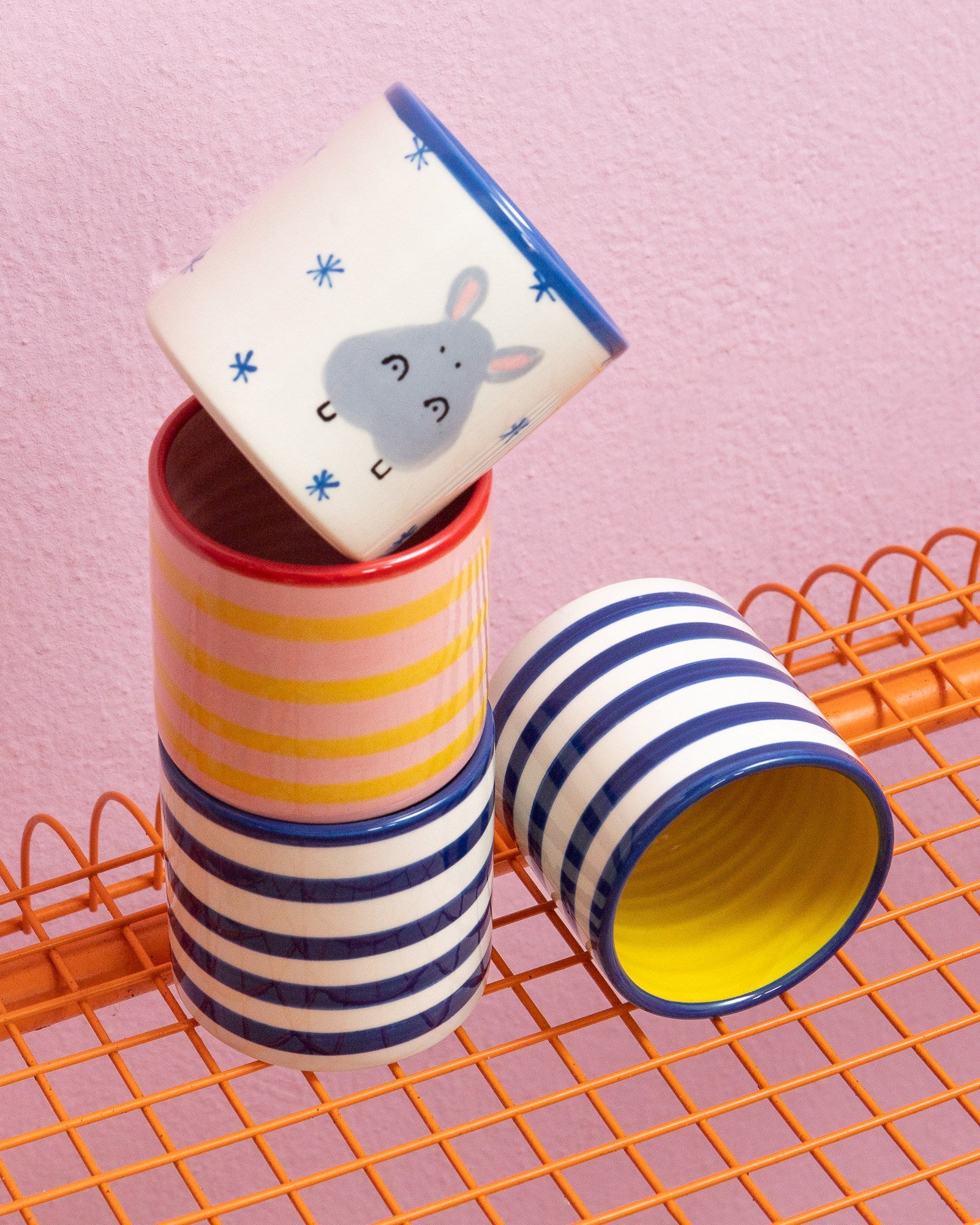 Vier bunte, gestreifte Becher und ein Becher mit Häschenmotiv auf einem orangen Gitterregal vor rosa Wand.