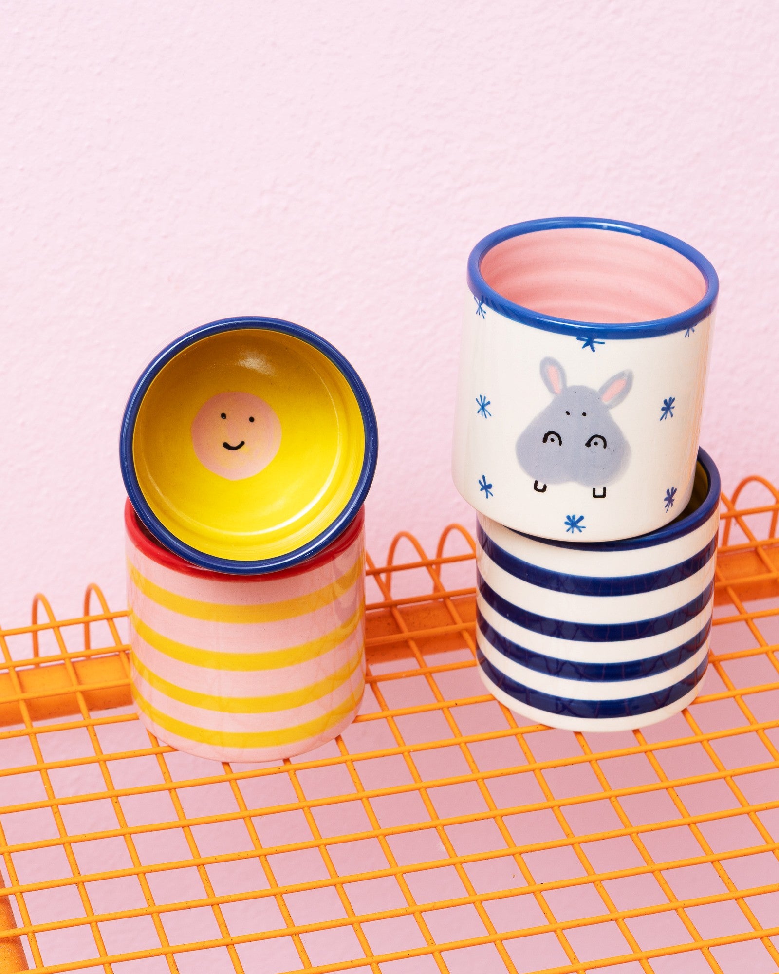 Mehrere bunt gestreifte Tassen mit fröhlichen Gesichtern und Tiermotiven stehen auf einem orangen Gitterregal vor einer rosa Wand.