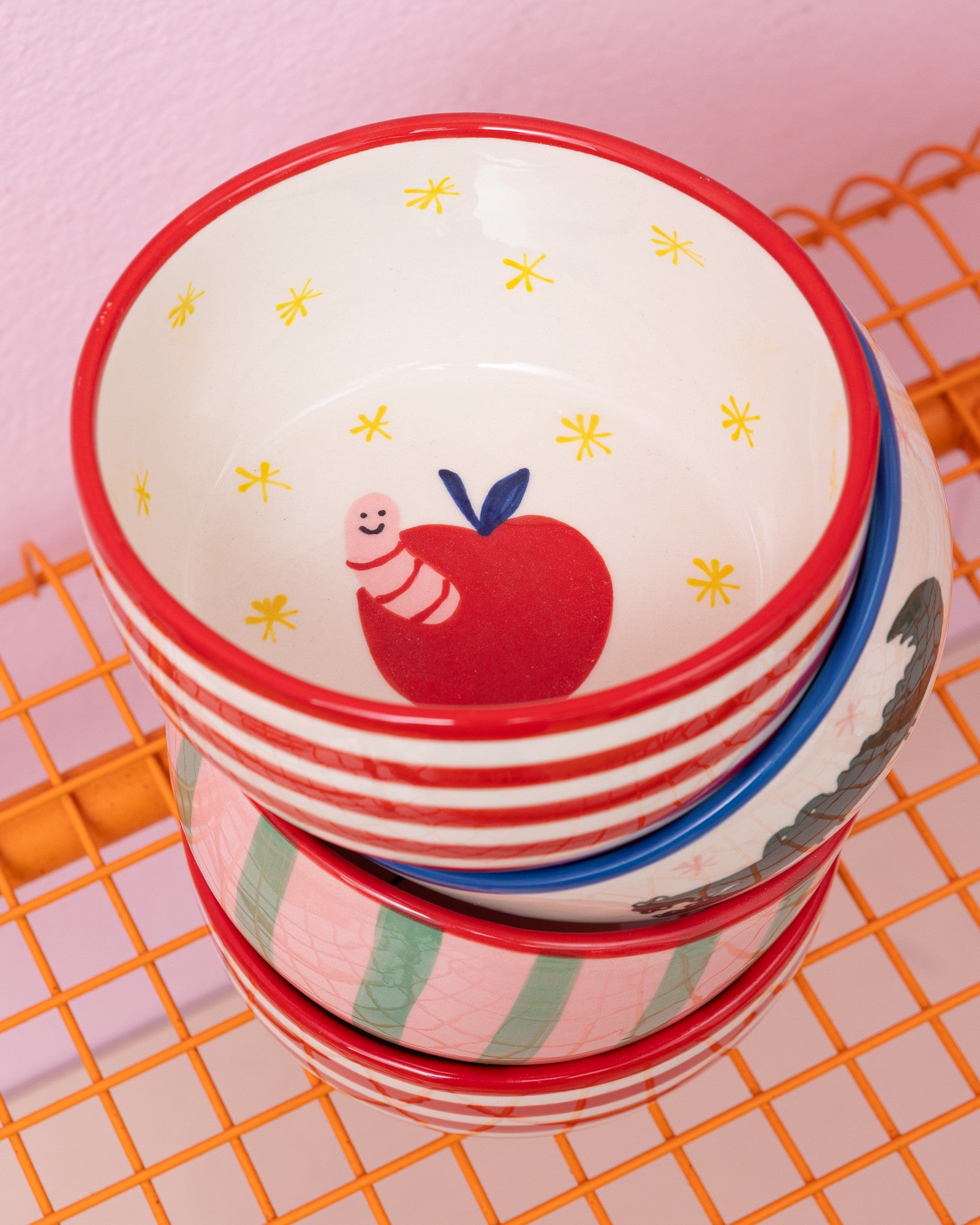 Ein Stapel bunter Schalen mit verschiedenen Mustern, darunter ein Apfel mit einem Wurm, auf einem orangefarbenen Gitter.