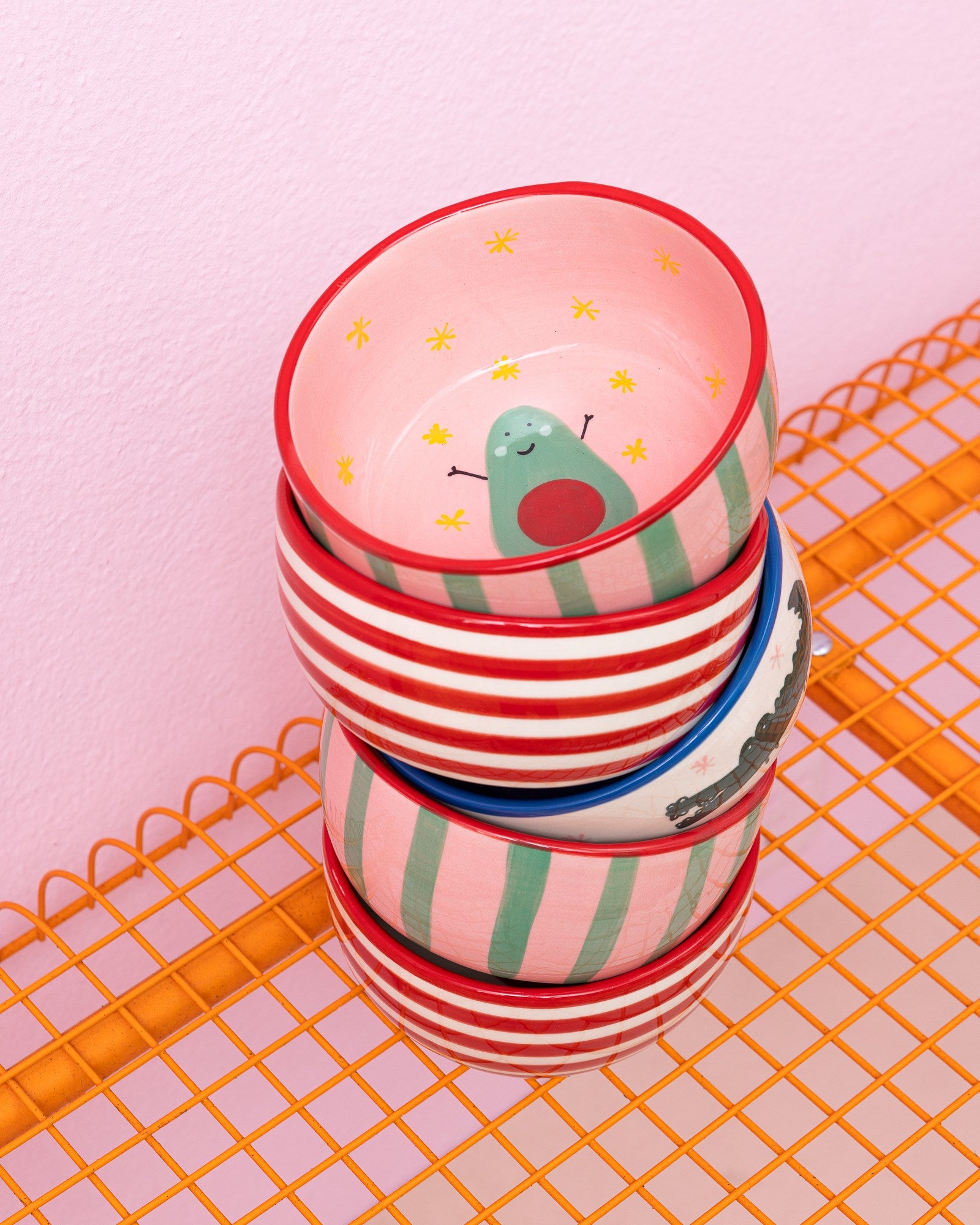 Ein Stapel bunt gestreifter Schüsseln steht auf einem orangefarbenen Gitterregal vor einer rosa Wand.