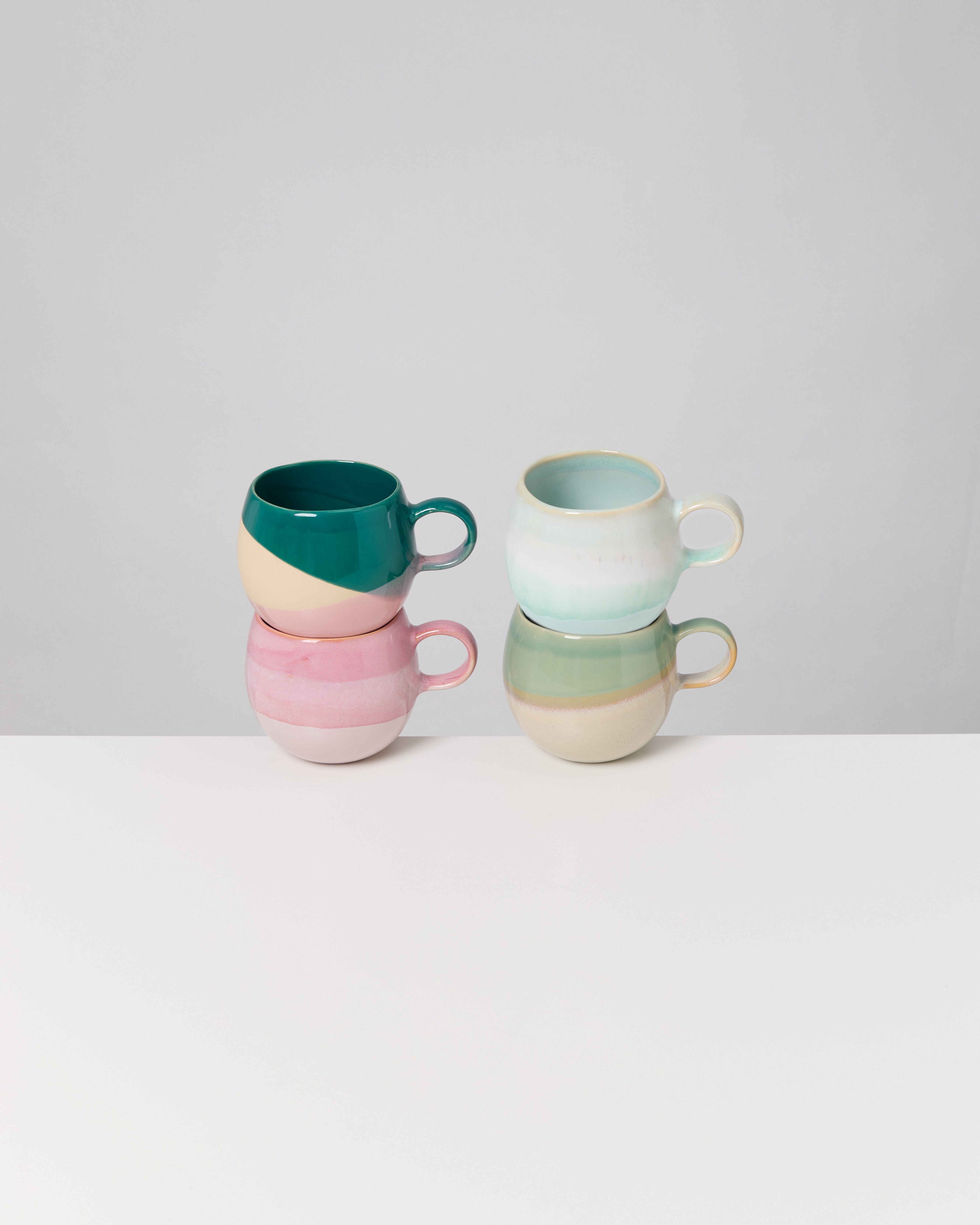 Vier bunte Tassen, zwei übereinander gestapelt, auf einer weißen Oberfläche vor grauem Hintergrund.