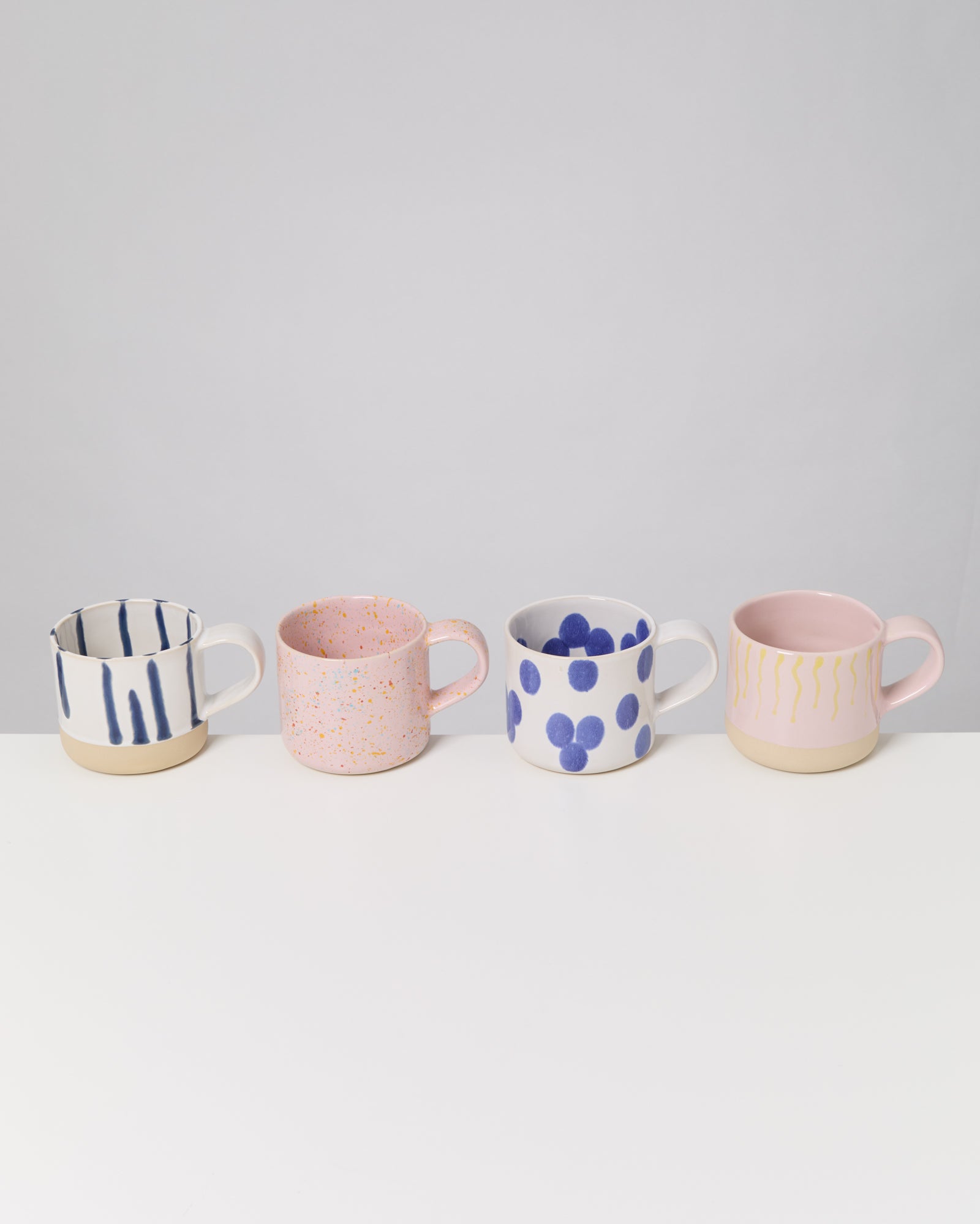 Vier bunte Tassen mit unterschiedlichen Mustern auf einer weißen Oberfläche vor grauem Hintergrund.