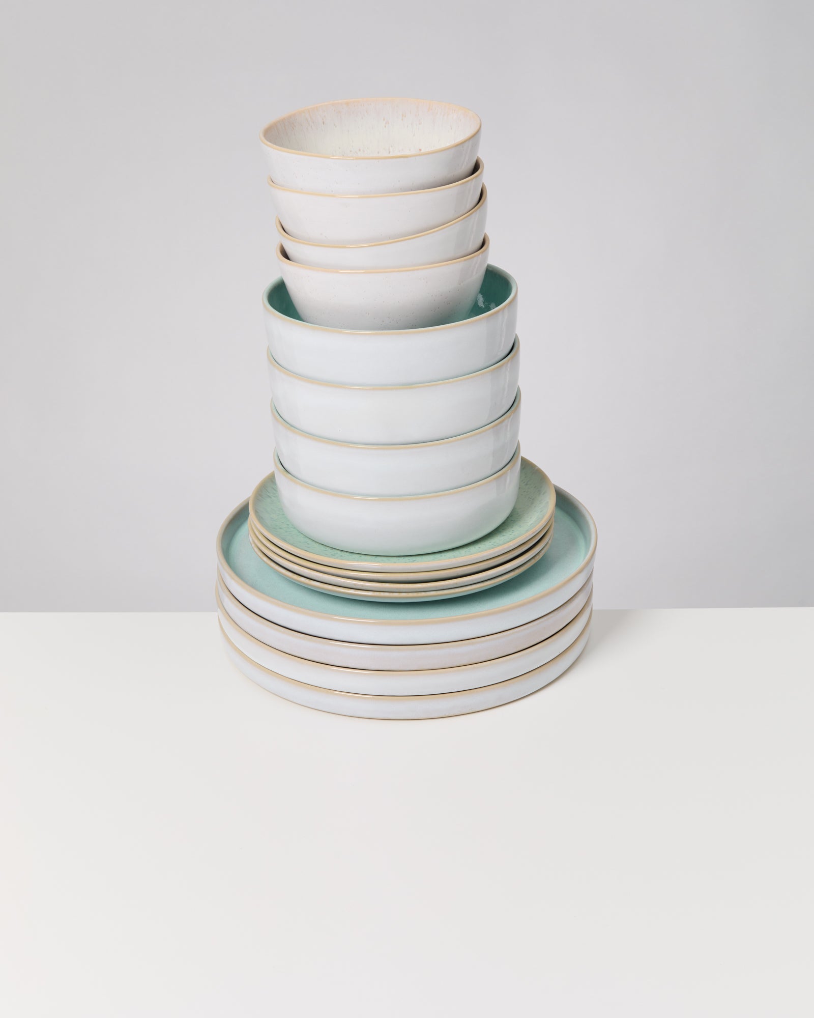 Ein gestapeltes Geschirrset mit hellen Schüsseln und Tellern auf einem weißen Tisch vor grauem Hintergrund.