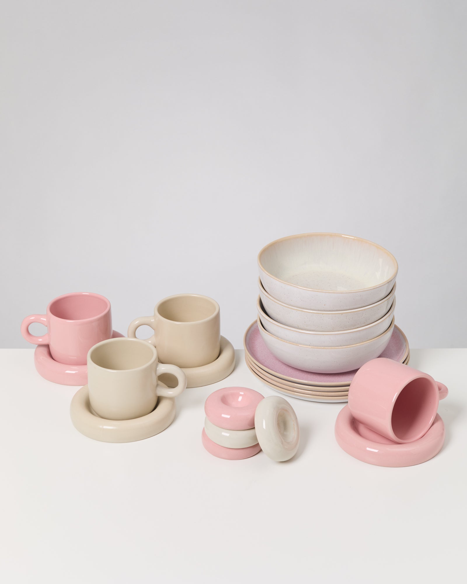 Rosafarbene und cremefarbene Tassen, Schüsseln und Untersetzer auf einem weißen Tisch angeordnet.