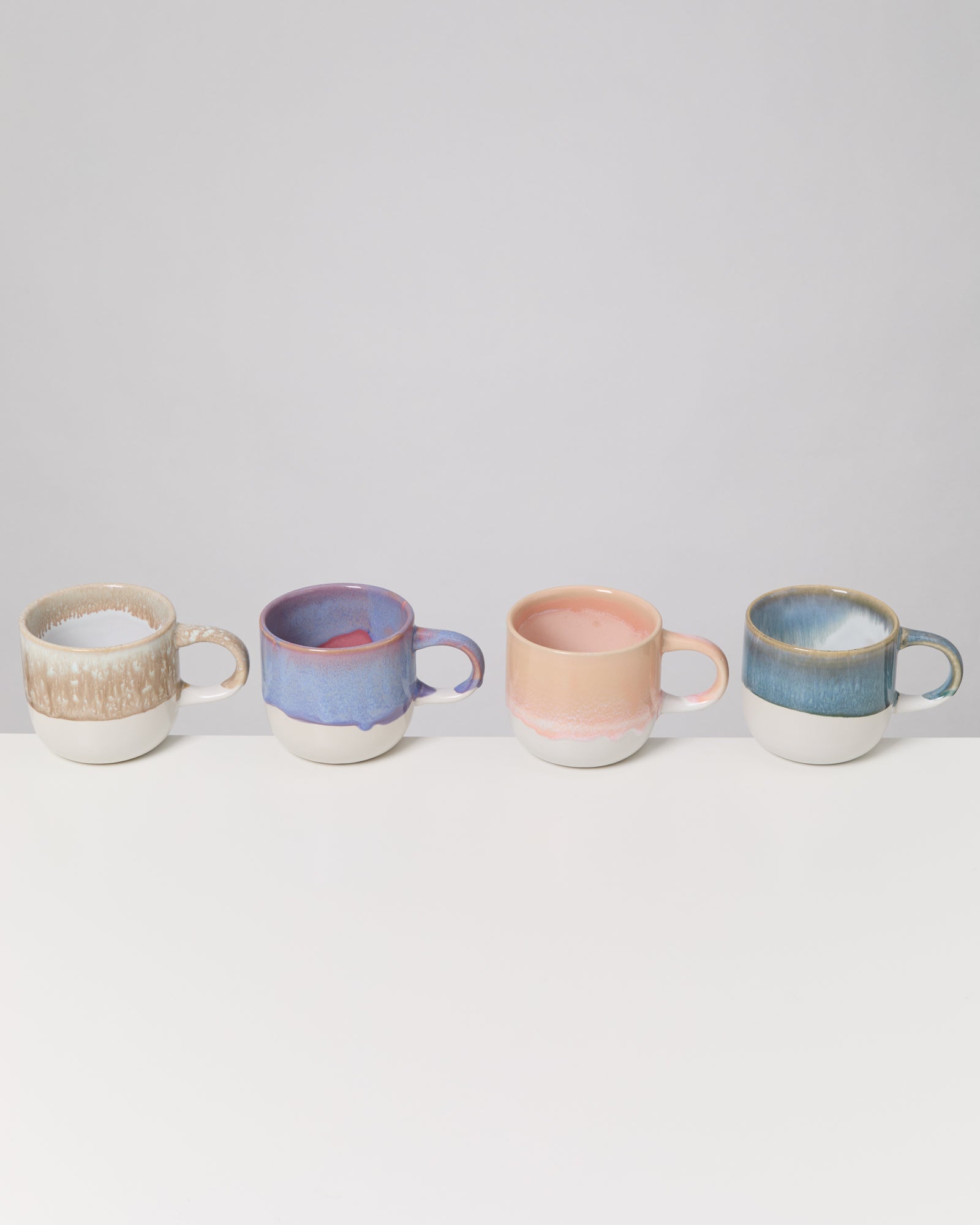 Vier unterschiedlich farbige Tassen stehen in einer Reihe auf einer weißen Oberfläche vor einem grauen Hintergrund.