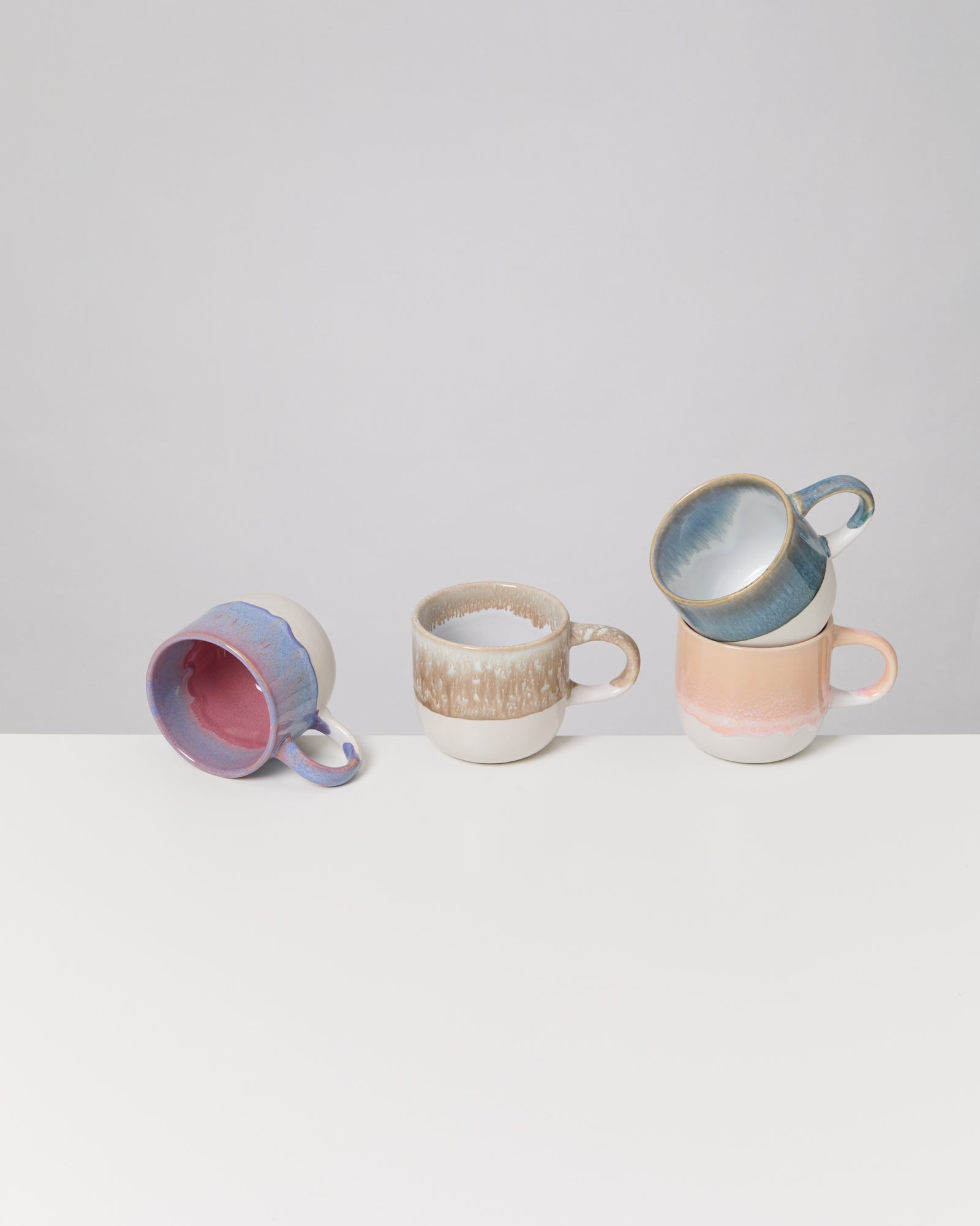 Vier bunte Keramiktassen stehen auf einem weißen Tisch, zwei übereinander gestapelt und eine auf der Seite liegend.