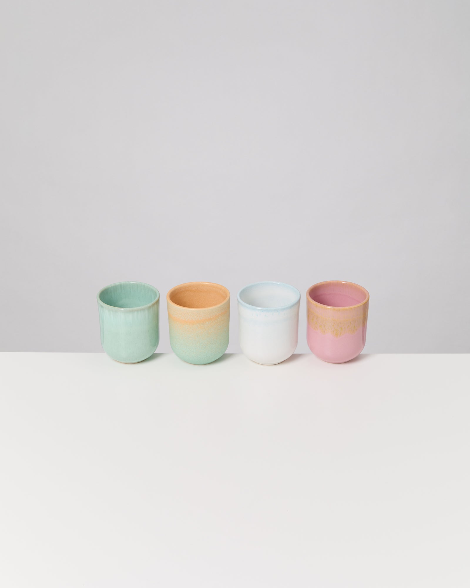 Vier bunte Keramiktassen stehen in einer Reihe auf einem weißen Tisch vor grauem Hintergrund.