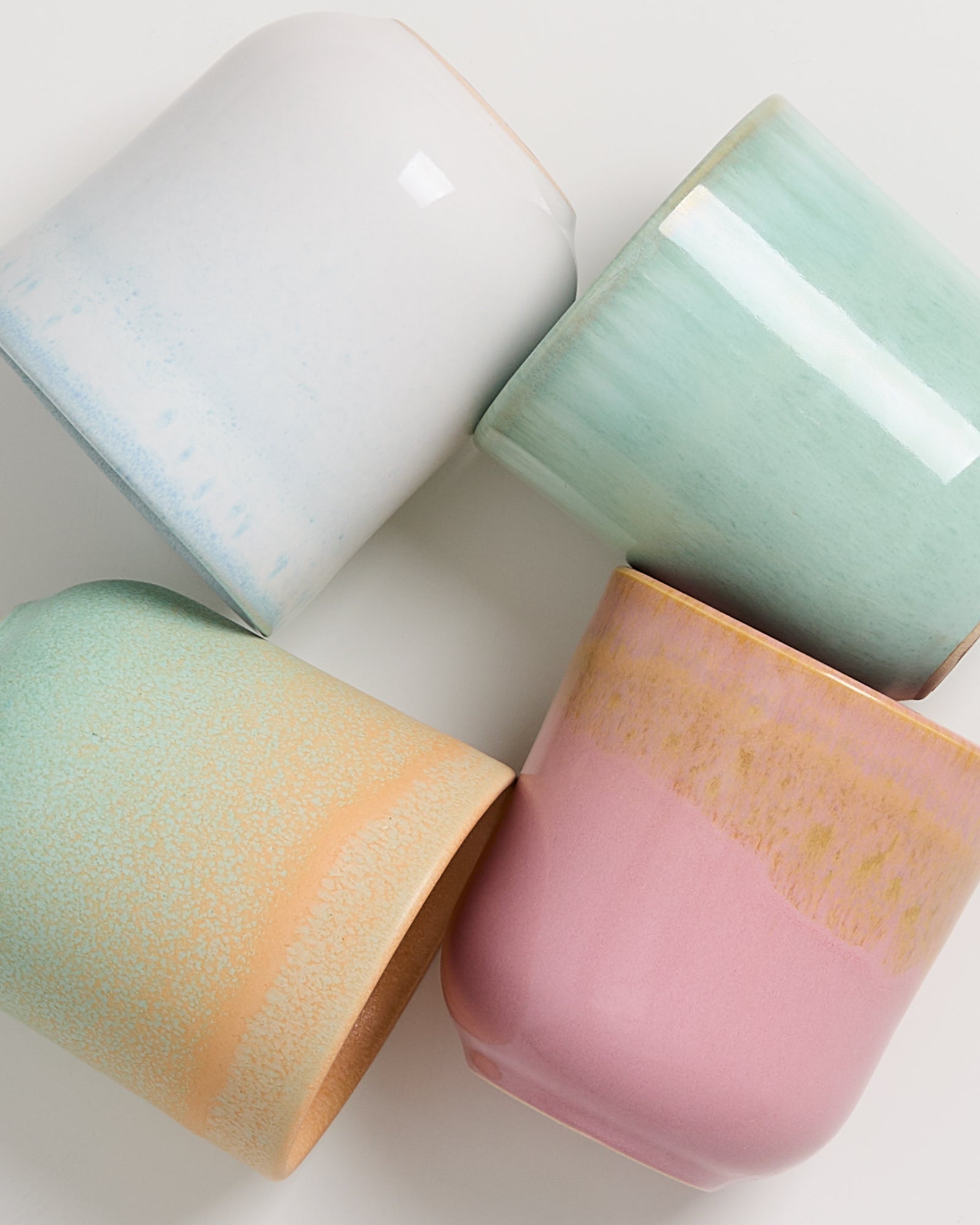 Vier bunte Keramiktassen in Pastelltönen liegen kreuzweise auf einer weißen Oberfläche.