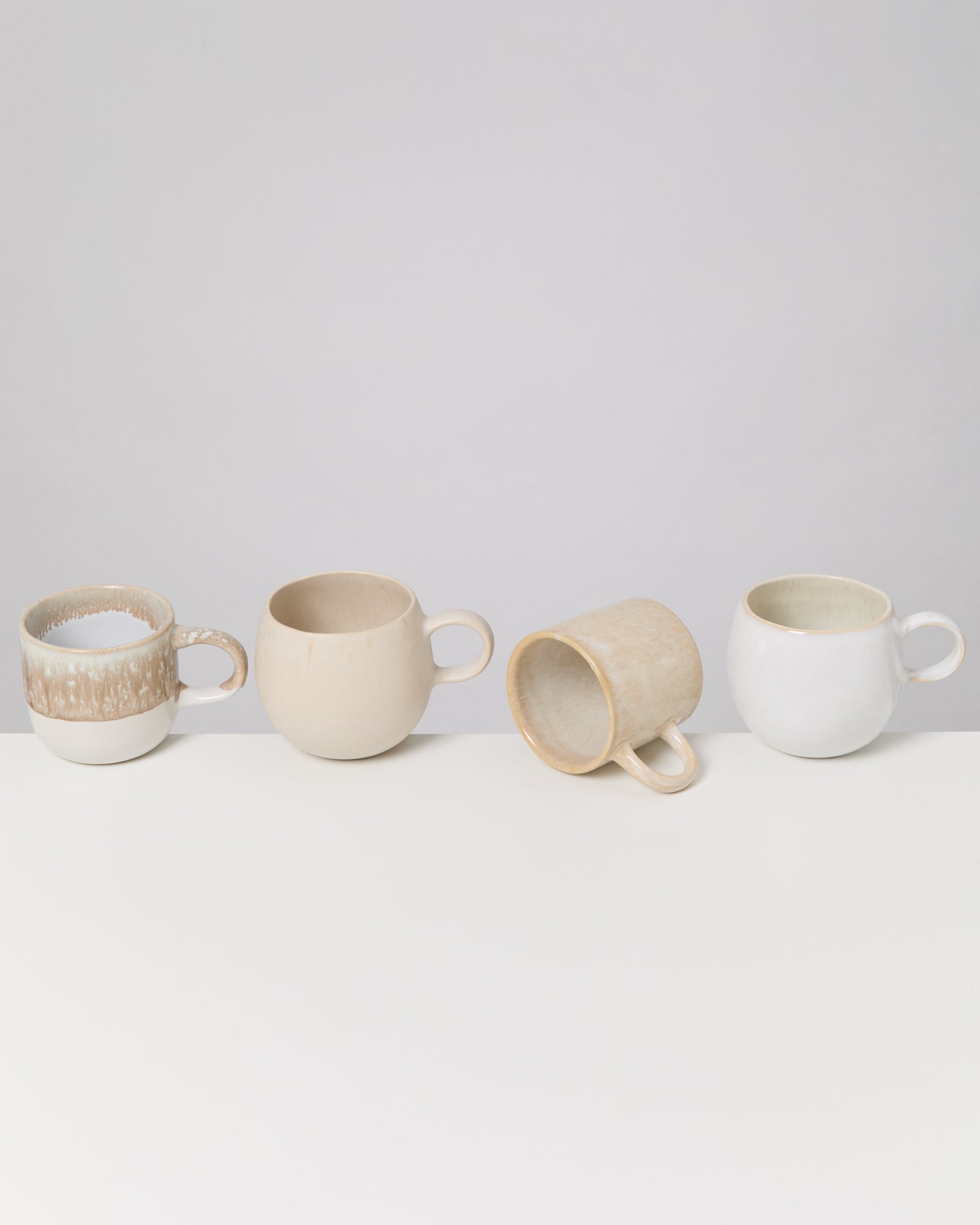 Vier hellfarbene Tassen mit Henkeln stehen nebeneinander auf einer weißen Oberfläche.