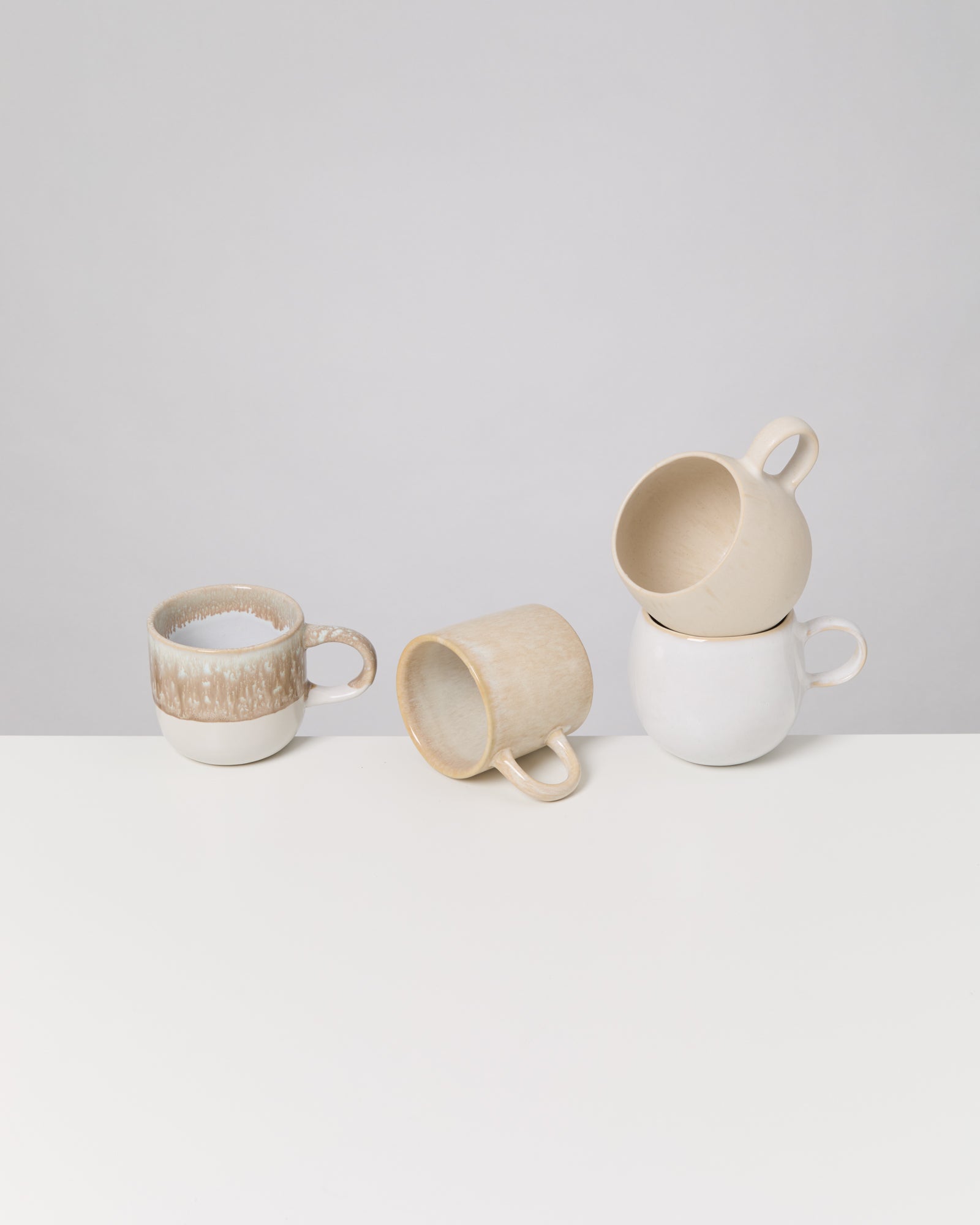 Vier Keramiktassen in neutralen Farbtönen auf einem weißen Tisch, zwei liegen, zwei sind gestapelt.