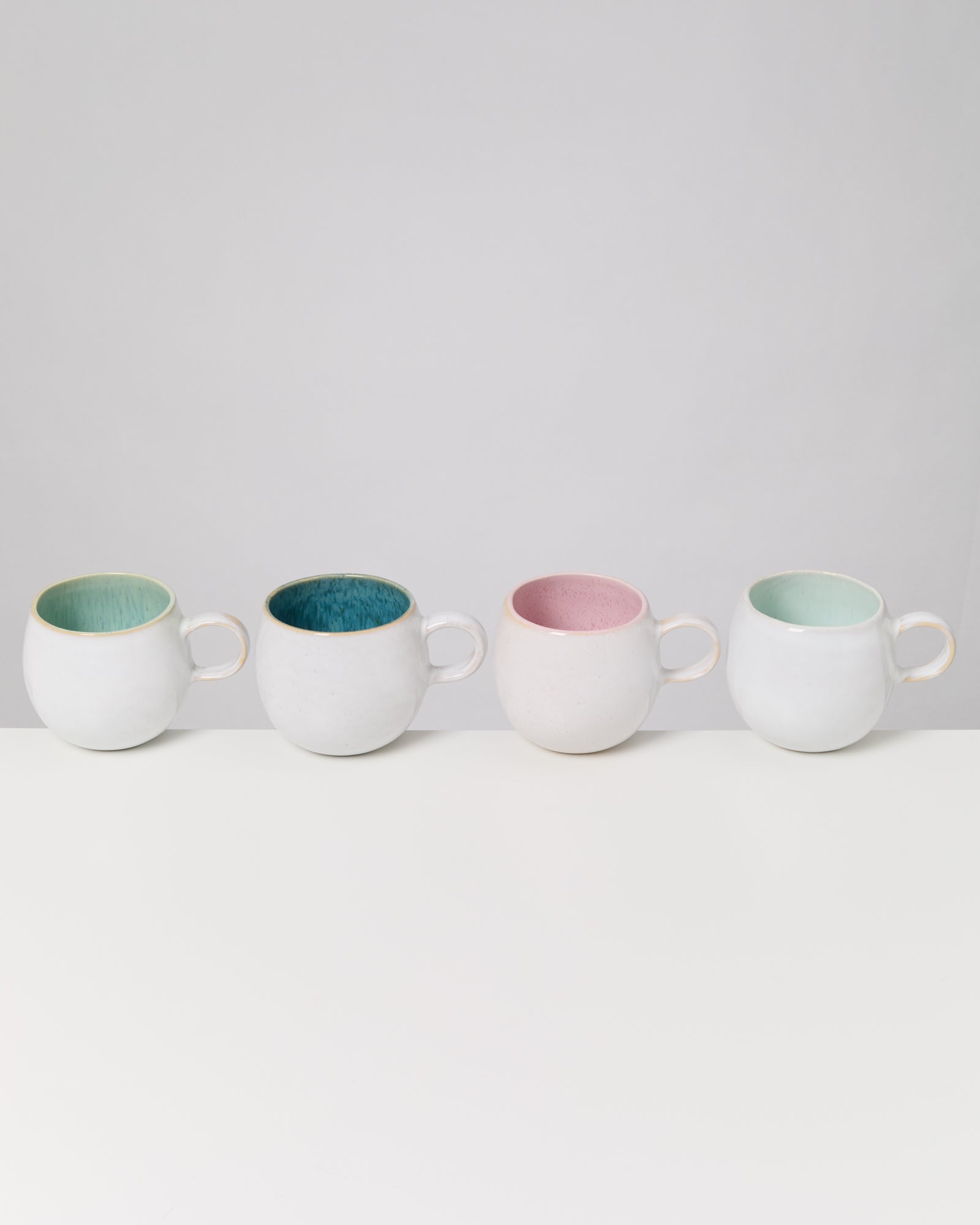Vier weiße Tassen mit verschiedenfarbigen Innenseiten (grün, blau, rosa, grün) stehen auf einem weißen Tisch.