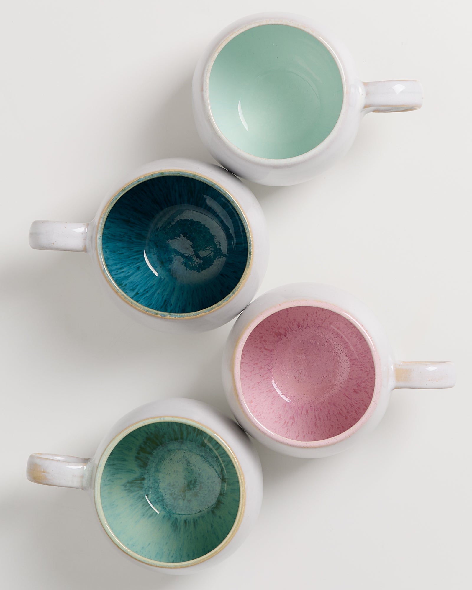 Vier Tassen in Weiß mit farbigem Innenleben in Mintgrün, Blau, Rosa und Grün auf einer weißen Oberfläche.