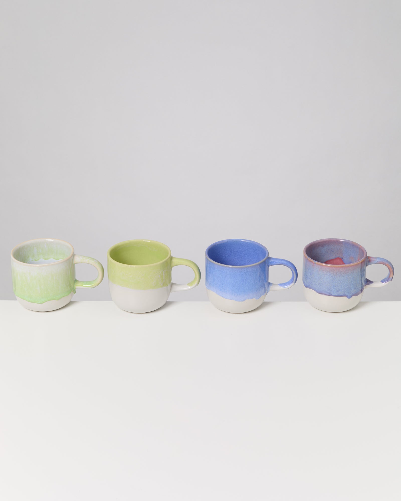 Vier bunte Keramiktassen in Grün, Blau und Violett stehen auf einem weißen Tisch vor grauem Hintergrund.