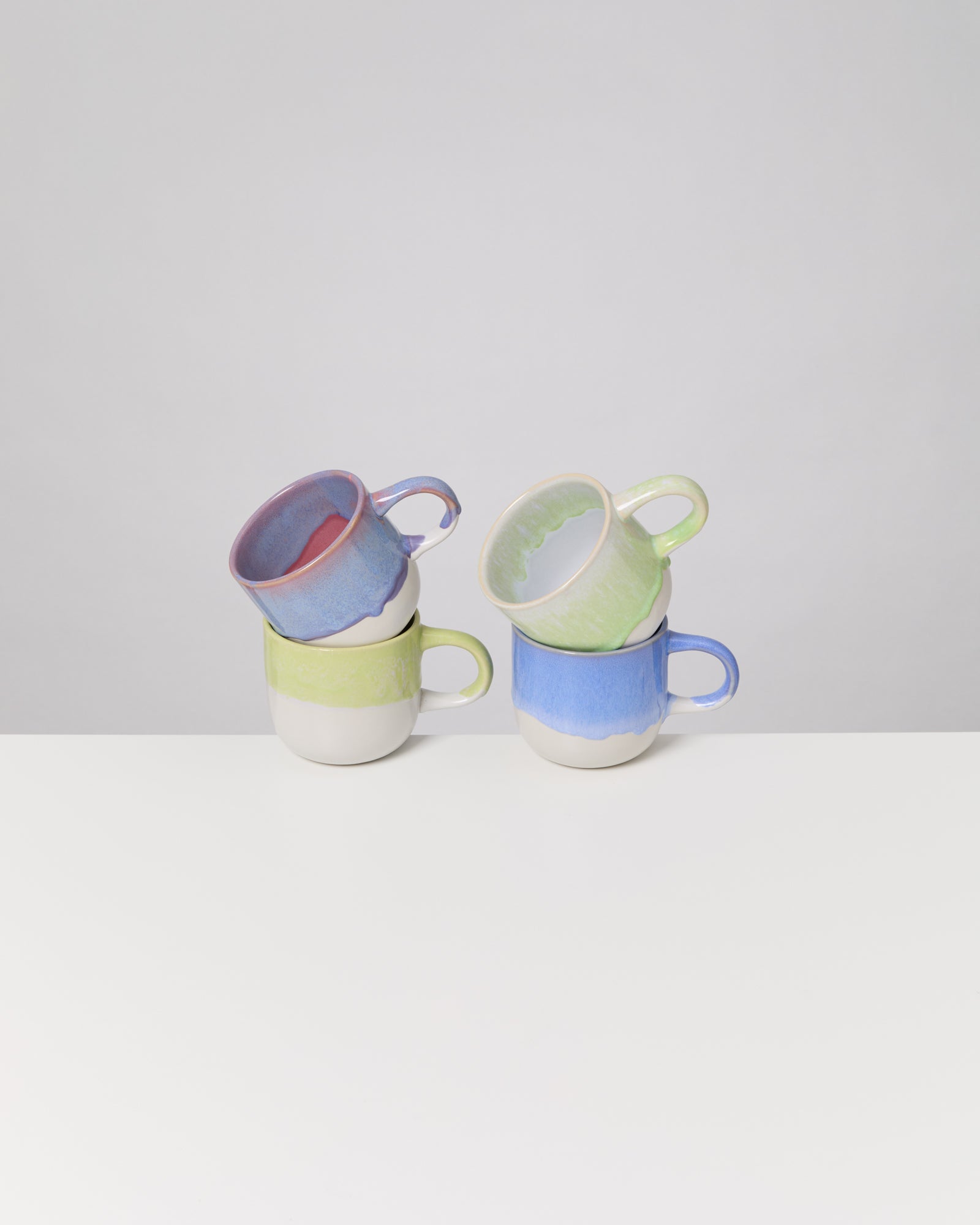 Zwei Stapel farbiger Keramiktassen stehen auf einem weißen Tisch vor grauem Hintergrund.
